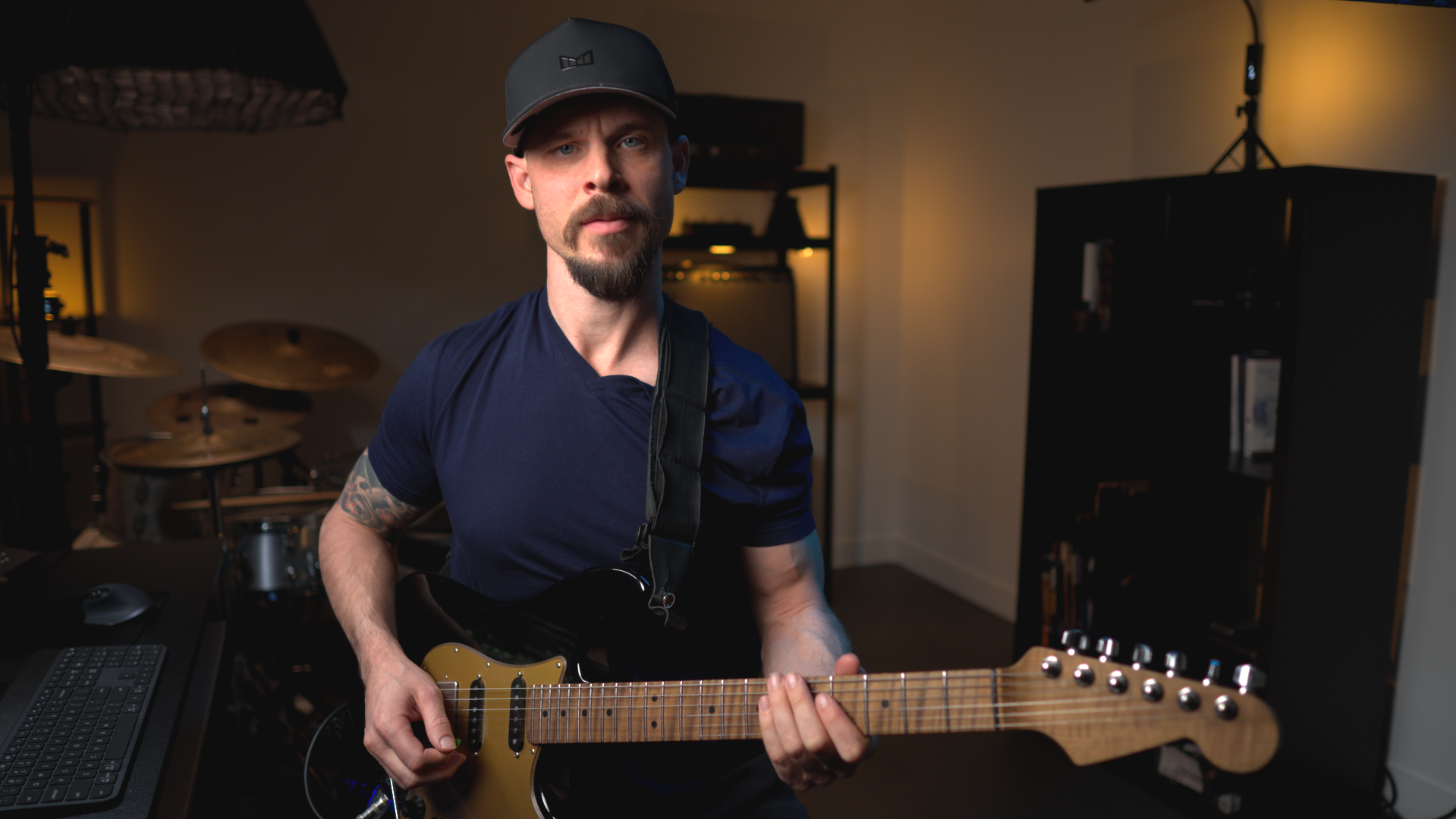 A man wearing a black cap and navy blue t-shirt playing an electric guitar in a music studio with drums and shelving units in the background.