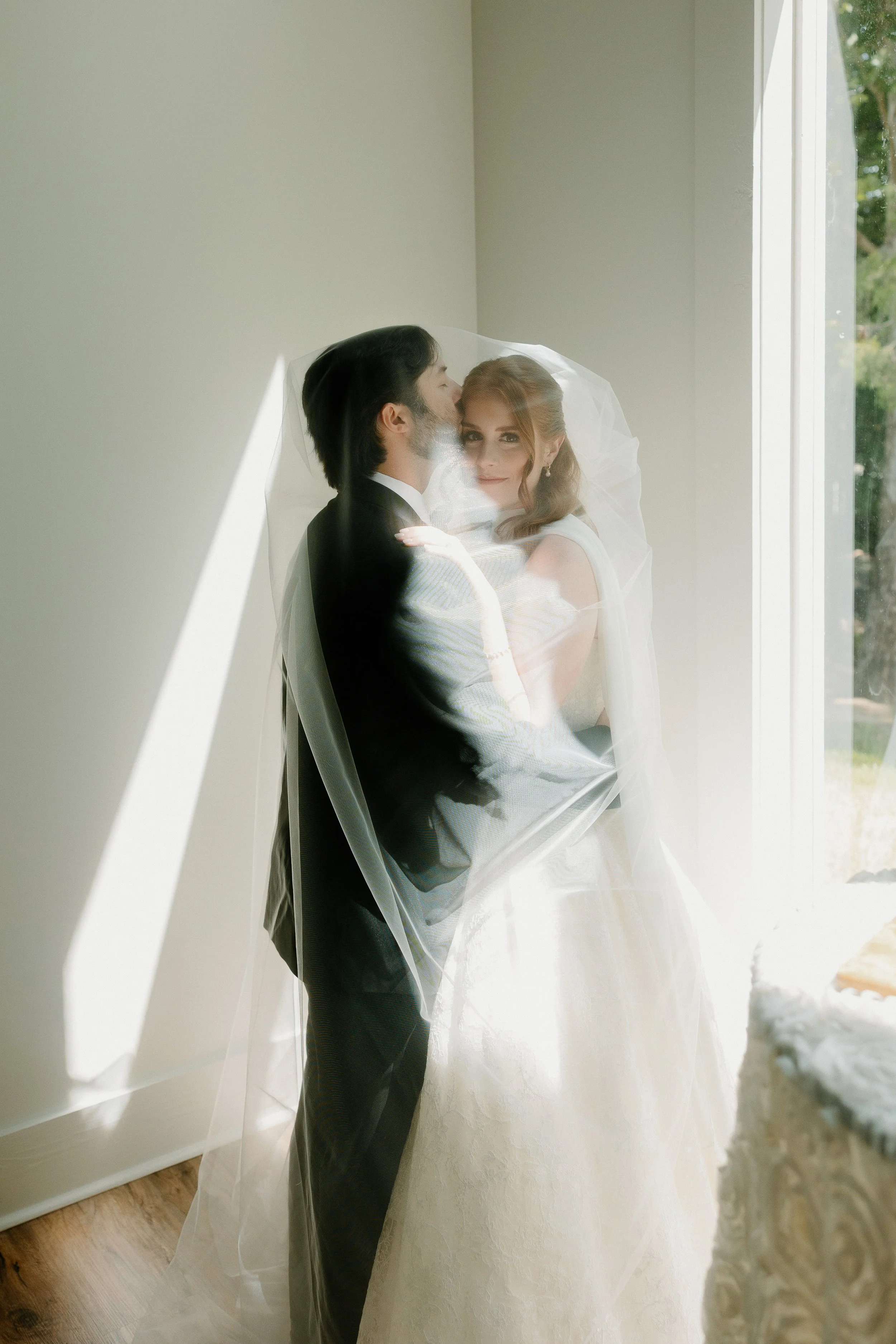 Bride and groom in wedding attire standing by a window, with the groom kissing the bride on the forehead, inside chapel at Kingfisher Lodge in Tupelo, Mississippi.