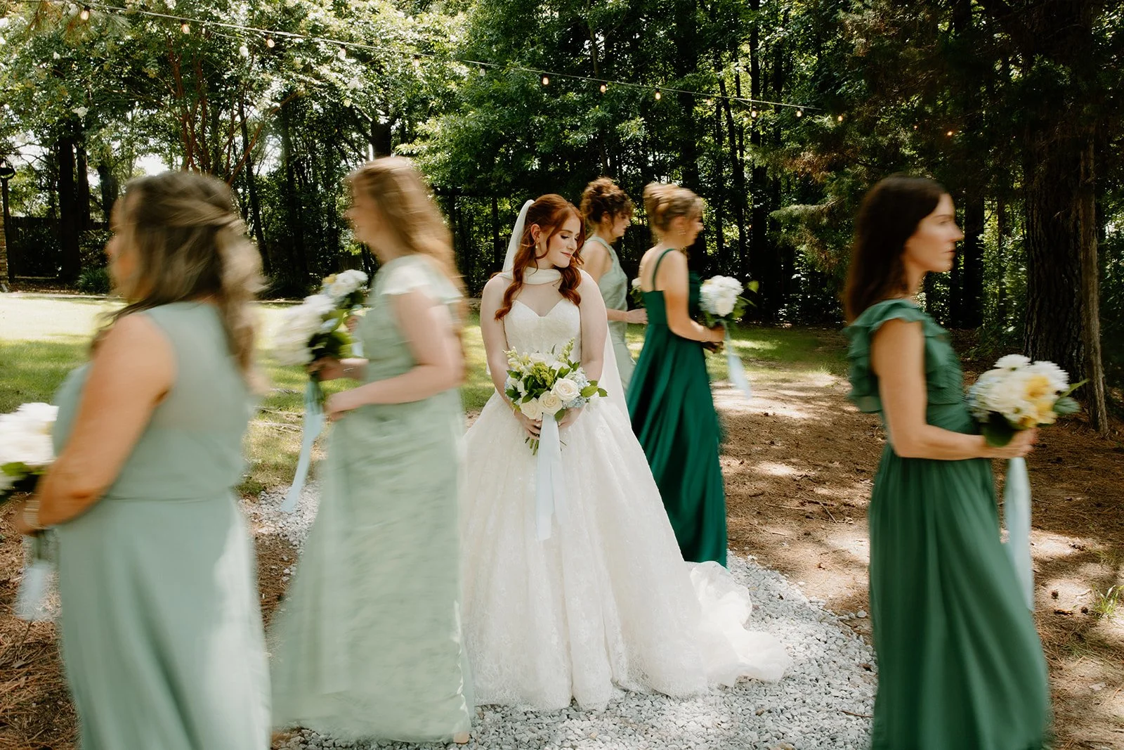 A bride in a white wedding dress holding a bouquet standing among bridesmaids in green dresses holding bouquets at Kingfisher Lodge venue on outdoor grounds. 