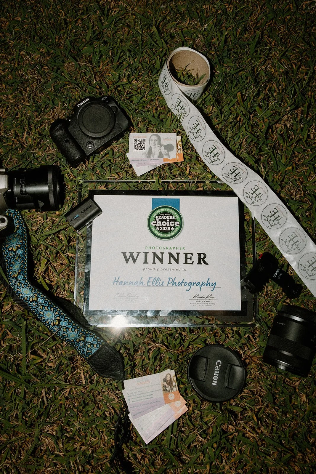 A photography award display on grass featuring a framed WINNER certificate for reader's choice award in Lee county, a Canon camera, a camera lens, a lens cap, business cards, a wrist strap, and a roll of stickers with a logo.