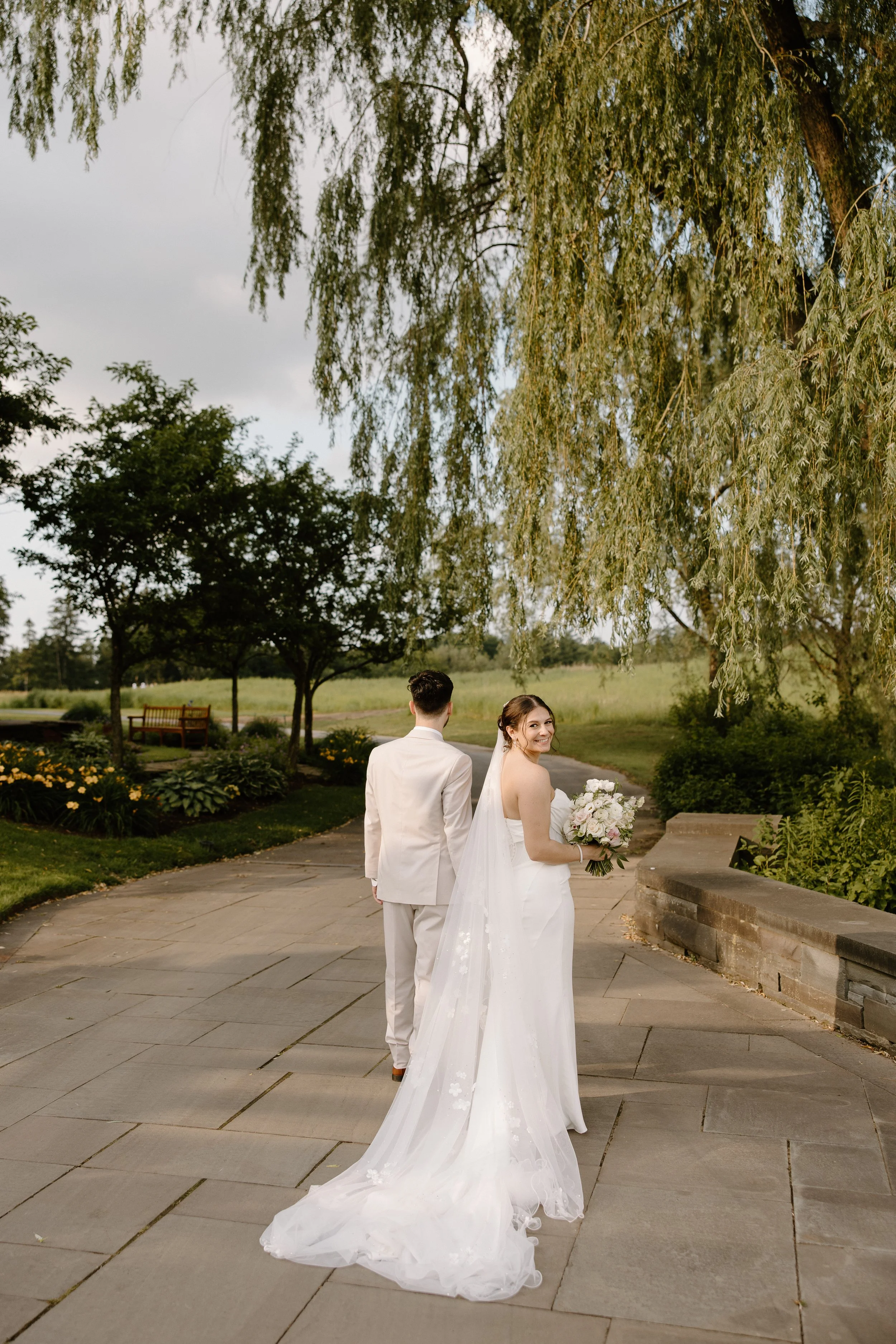 Bride in a white wedding dress holding a bouquet, smiling and looking back over her shoulder, with a groom in a white suit walking ahead on a paved path in a park.
