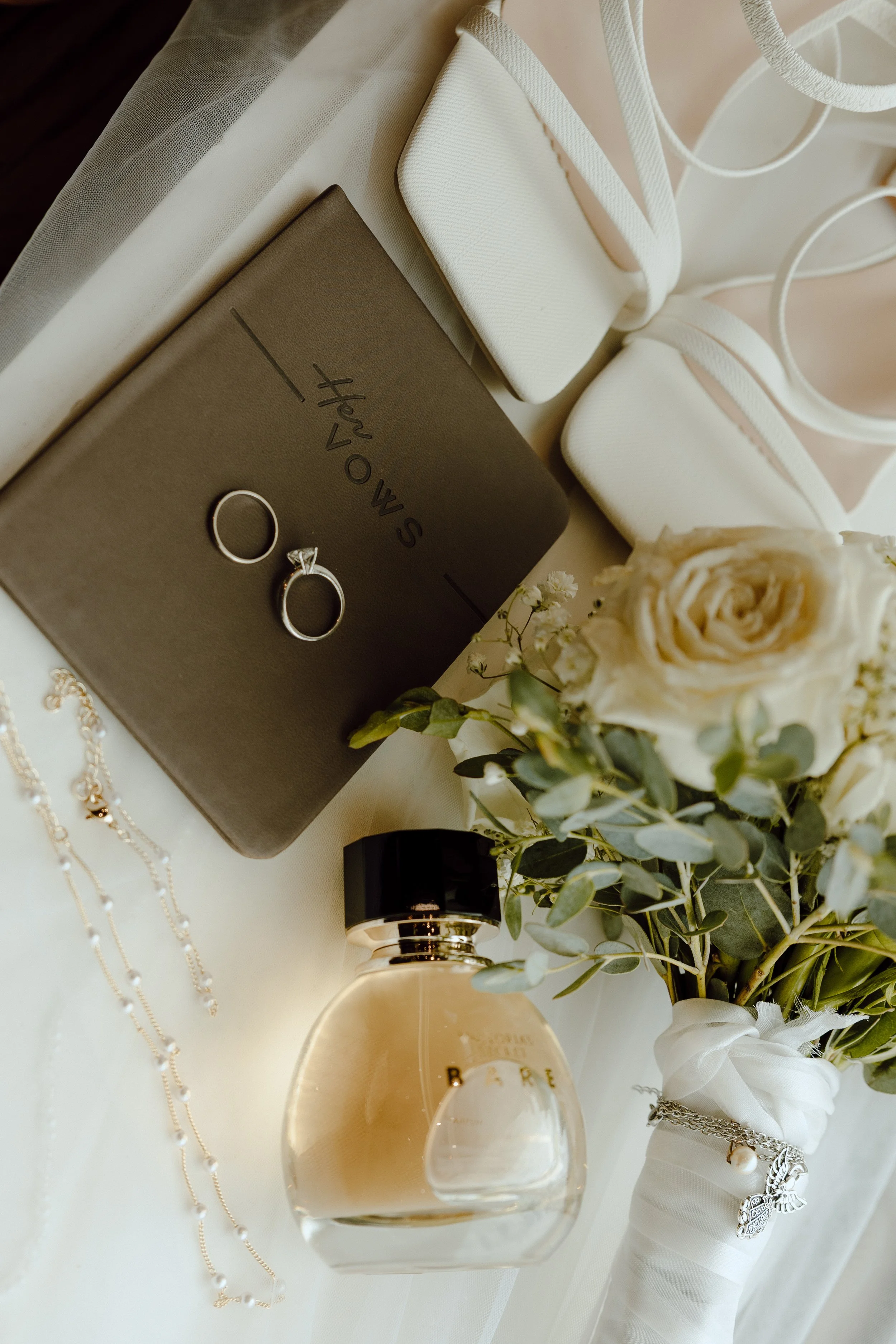 Wedding accessories including rings, jewelry, perfume bottle, flower bouquet, and wedding shoes arranged on a table.