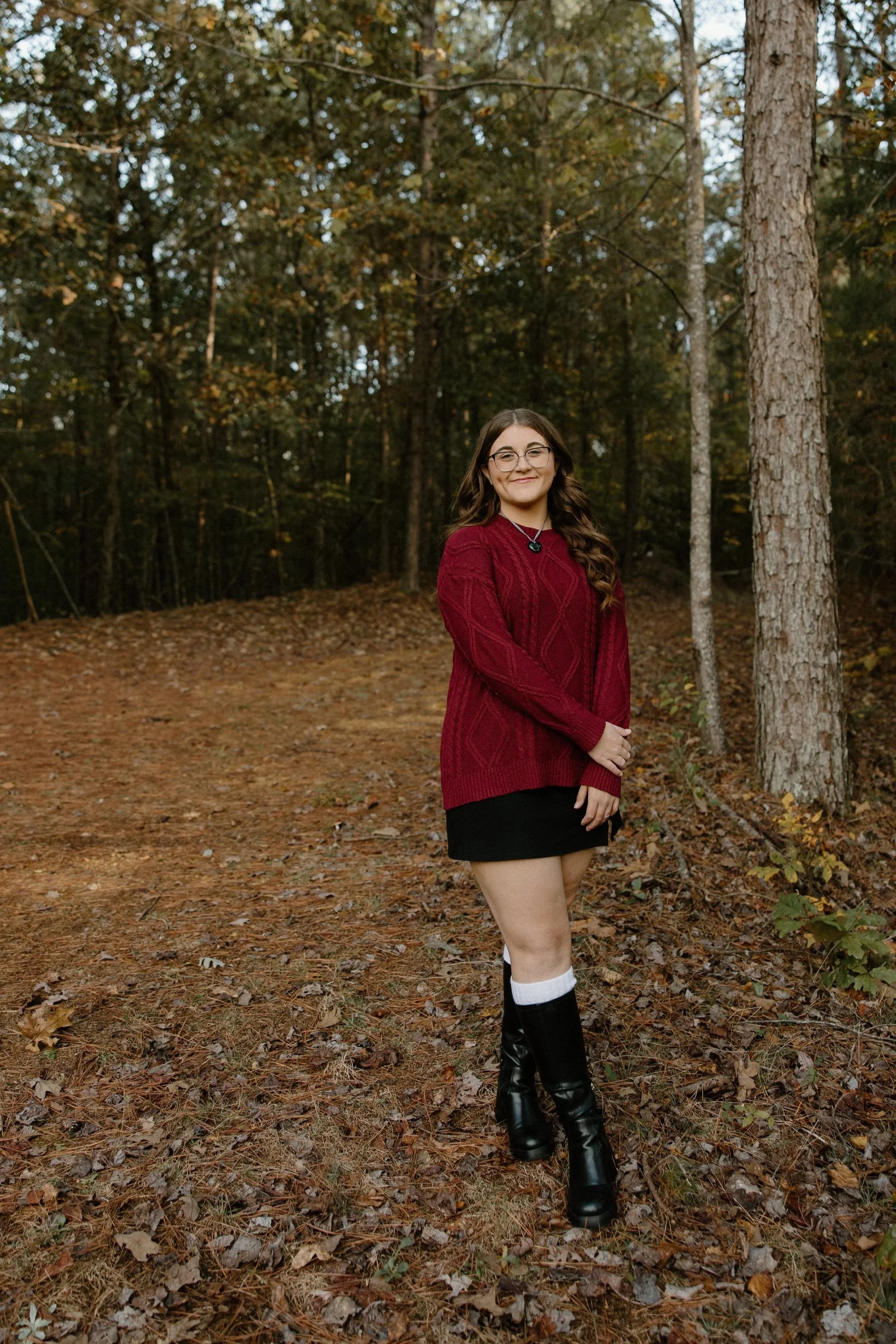 A young woman standing on a forest trail during fall, wearing a red sweater, black skirt, knee-high black boots with white socks, and glasses, smiling at the camera.