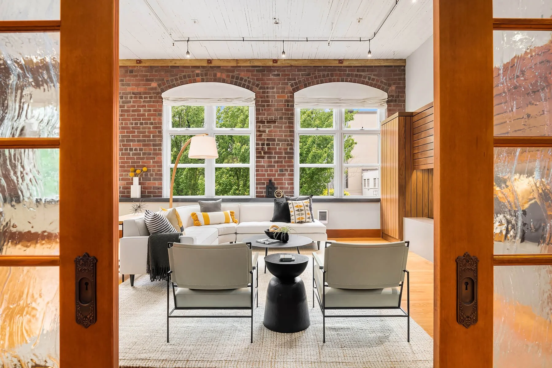 Living room with exposed brick wall, large windows showing green trees, white sectional sofa with decorative pillows, modern black side table, two white armchairs, and wooden accents.