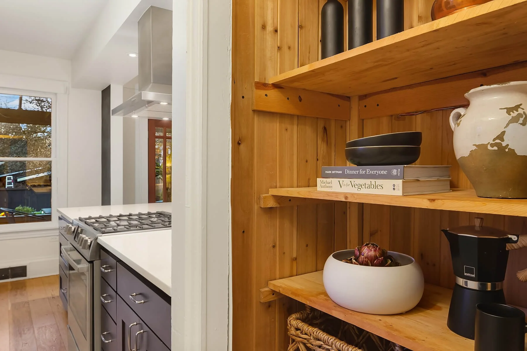 A kitchen interior with a white stove, stainless steel range hood, window, and a wooden shelving unit with cookbooks, bowls, and decorative items.