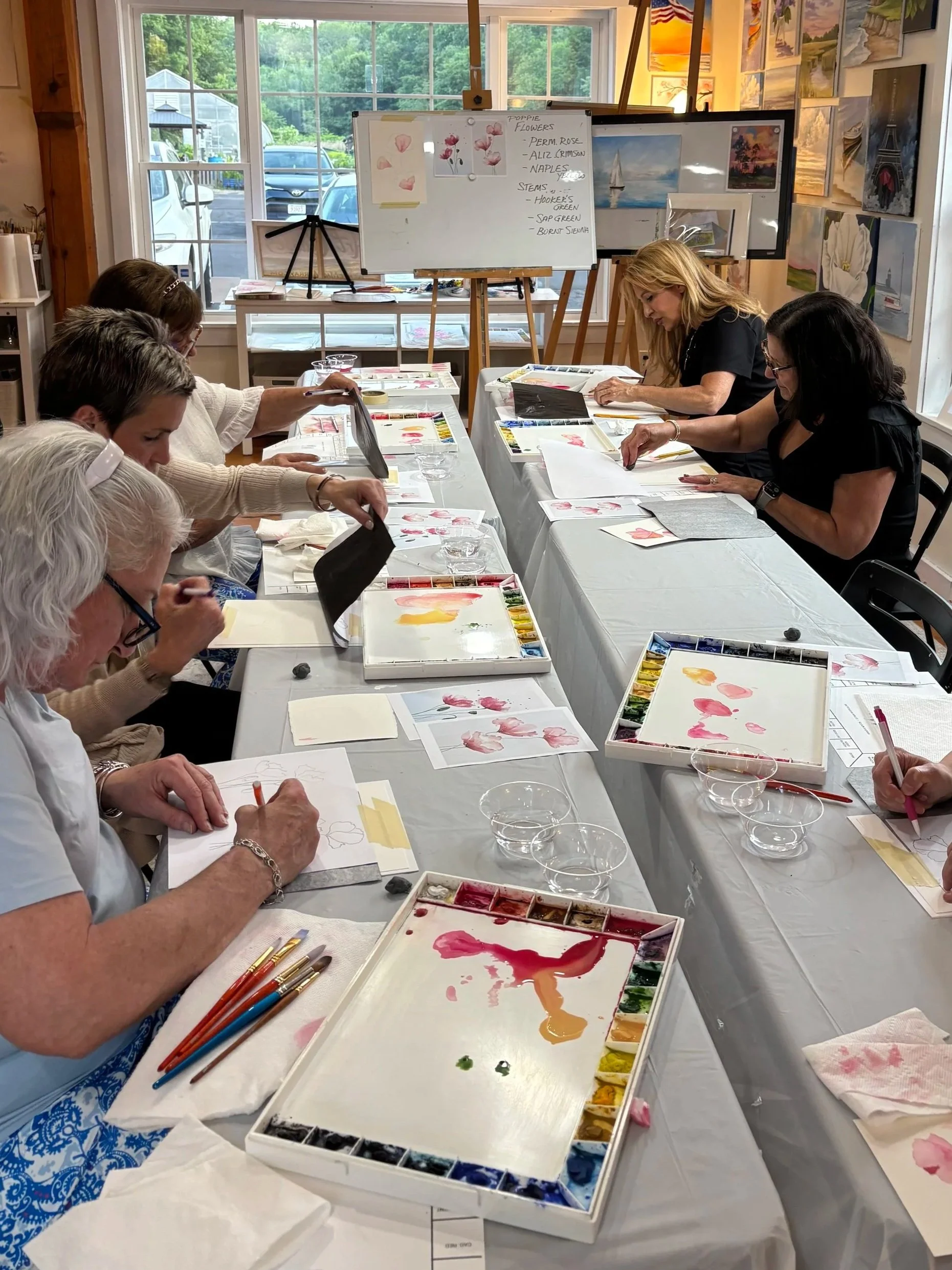 A group of people participating in a watercolor painting class in a well-lit room with large windows. They are working on watercolor paintings of pink flowers, with painting supplies and palettes on the table.