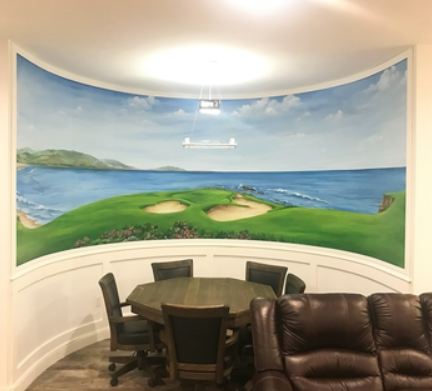 A mural of a seascape with green hills, sandy beaches, and blue ocean waves on a curved wall behind a round wooden table with five black chairs, in a room with a brown leather recliner and bright overhead lighting.