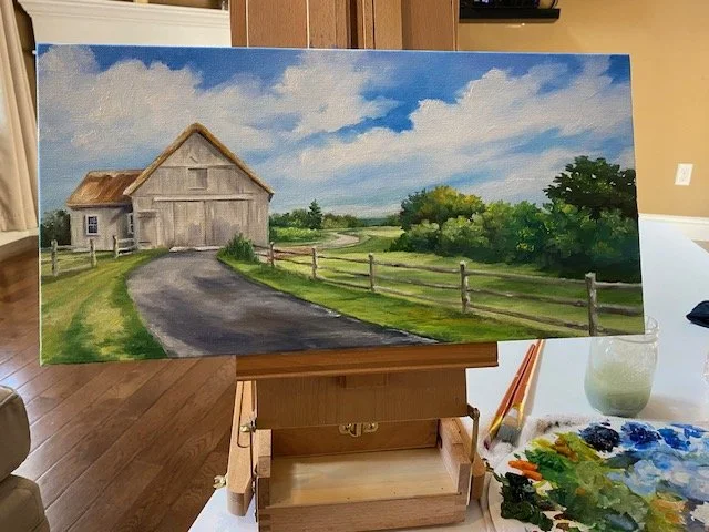 A landscape painting on an easel depicts a country scene with a barn, a winding road, green fields, trees, and a partly cloudy sky.