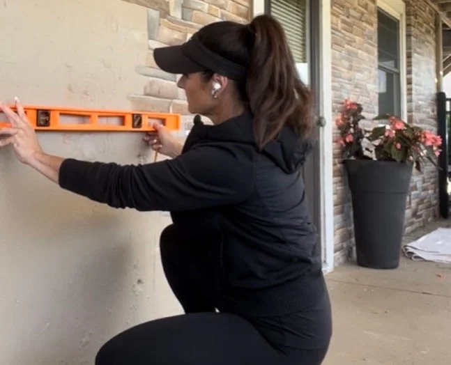 Woman using a spirit level to measure on an exterior wall of a house, wearing a black hoodie, black cap, and wireless earbuds, with a potted plant and building materials nearby.