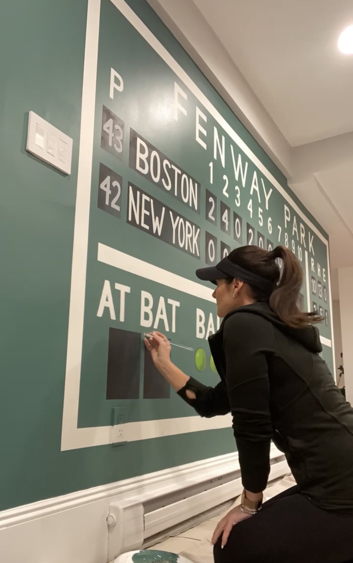 Woman with a black visor and hoodie writing on a large train schedule wall with a green marker.