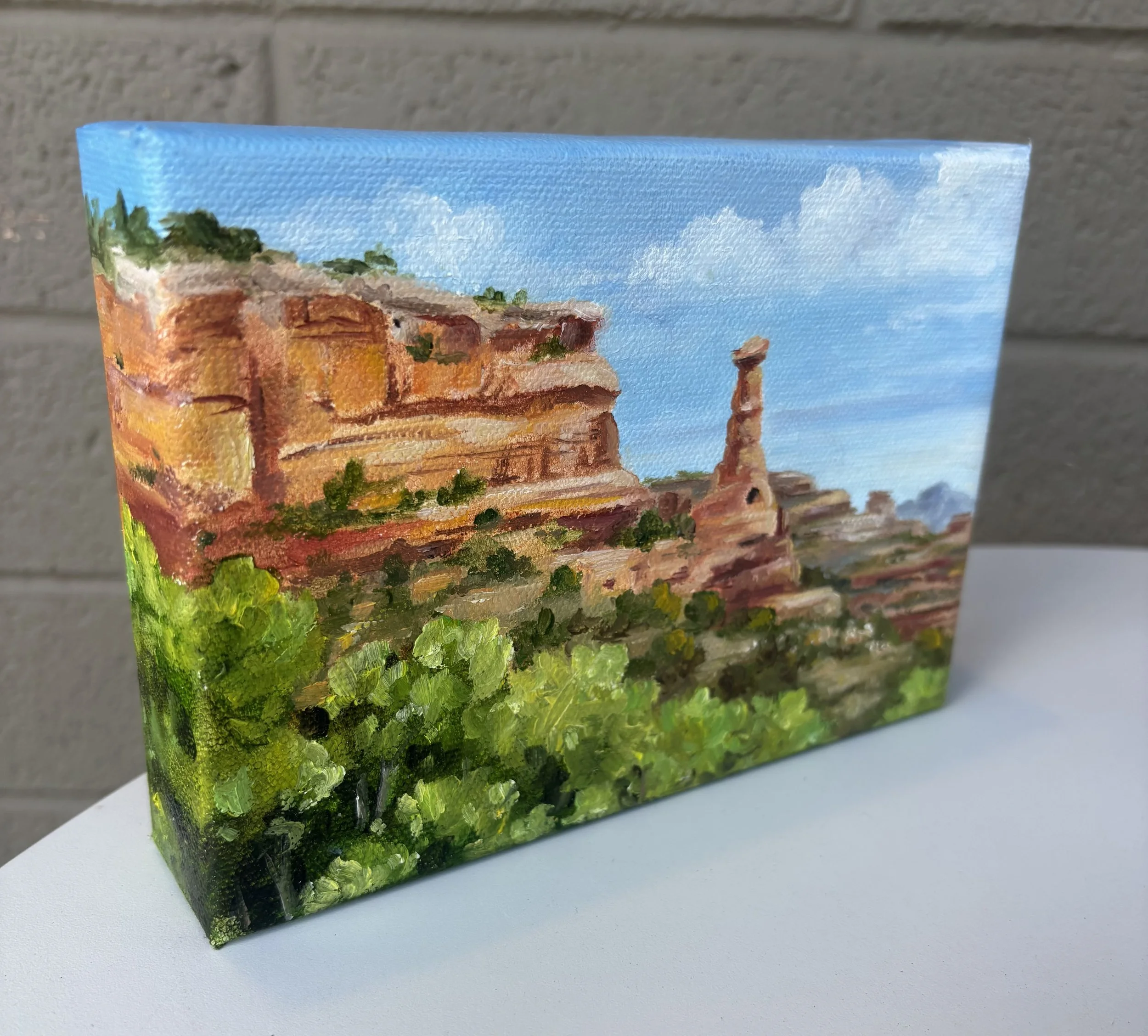 A painted wooden cube featuring a landscape scene with a blue sky, clouds, trees, and a red and yellow building, depicted with thick, textured brushstrokes.