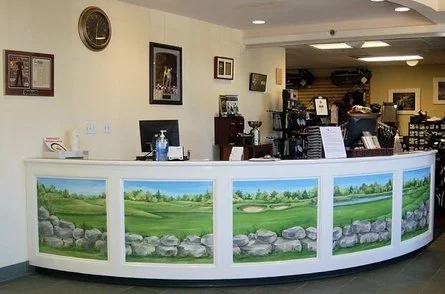 Interior of a reception area with a curved counter featuring landscape painting panels, framed pictures and plaques on the wall, and various items on the counter.