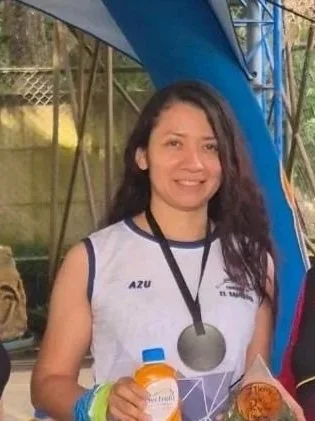 Young woman with long dark hair smiling, wearing a white sleeveless athletic shirt with blue trim, holding orange and yellow bottles, outdoors near a blue structure.