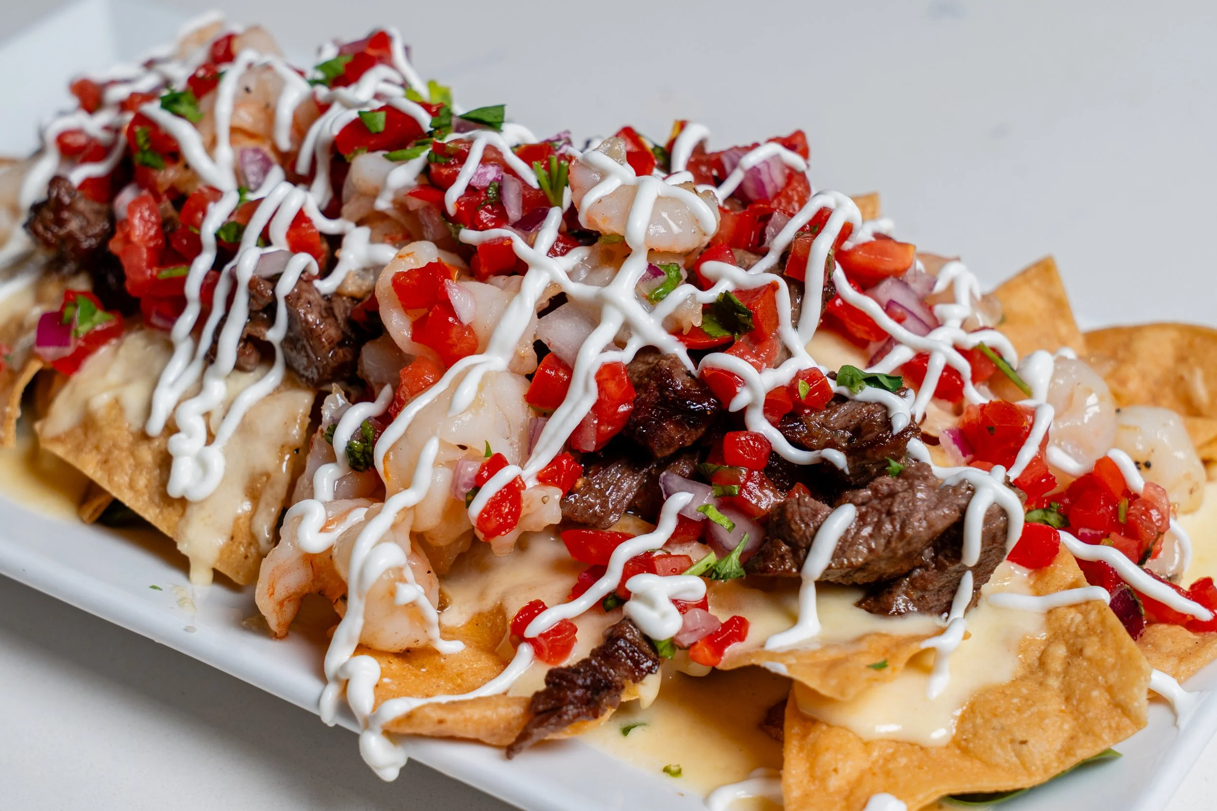 Close-up of a plate of loaded nachos topped with diced tomatoes, onions, cilantro, shredded meat, and drizzled with white sauce or sour cream.