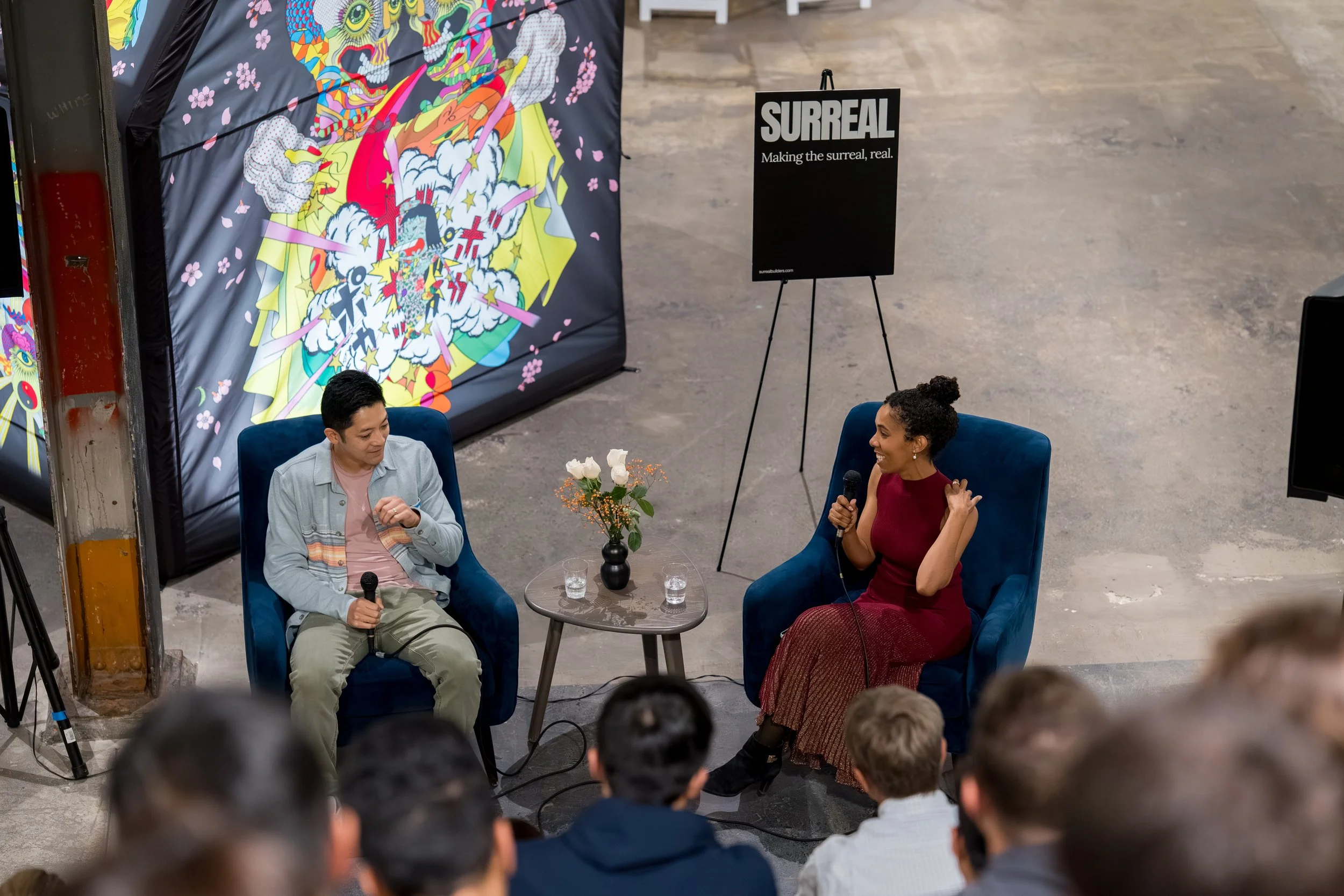 Two people sitting in blue armchairs engaging in conversation during an event. The man on the left holds a microphone, wears a light gray jacket, and has short dark hair. The woman on the right is smiling, holding a microphone, and wears a sleeveless red dress with curly hair styled in a bun. A small round table with a flower arrangement and two glasses is between them. Behind them, a colorful, comic-book style backdrop is displayed, and a black sign reads 'SURREAL Making the surreal, real.' An audience is visible in the foreground.