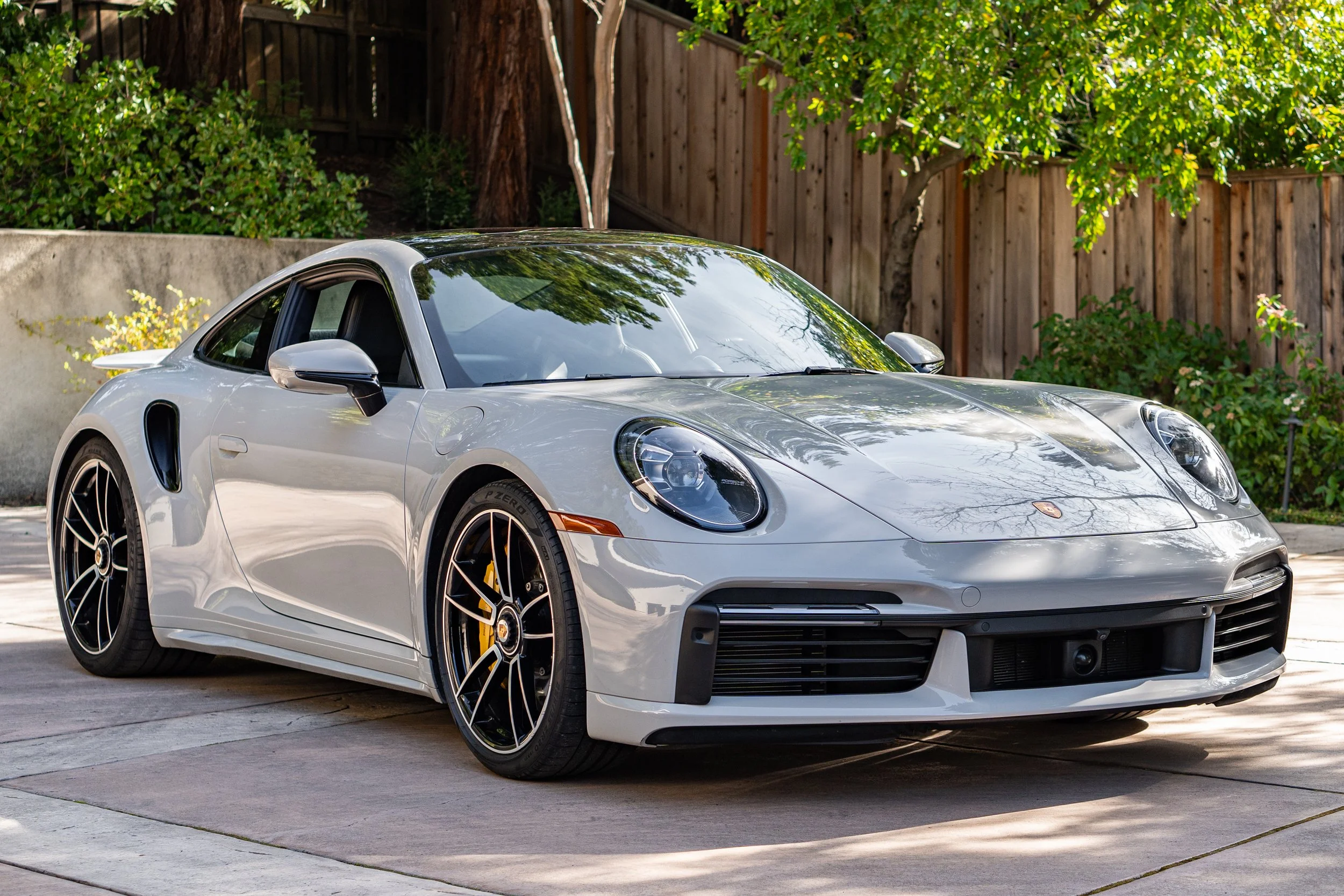 A silver Porsche sports car parked on a driveway with a wooden fence and green trees in the background.