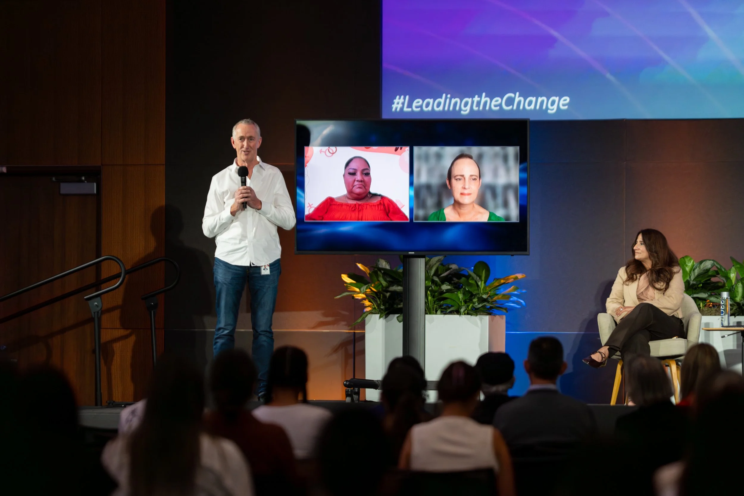 A man in a white shirt holding a microphone standing next to a large screen displaying two women in video call during a conference presentation with the hashtag #LeadingtheChange projected on the wall behind.