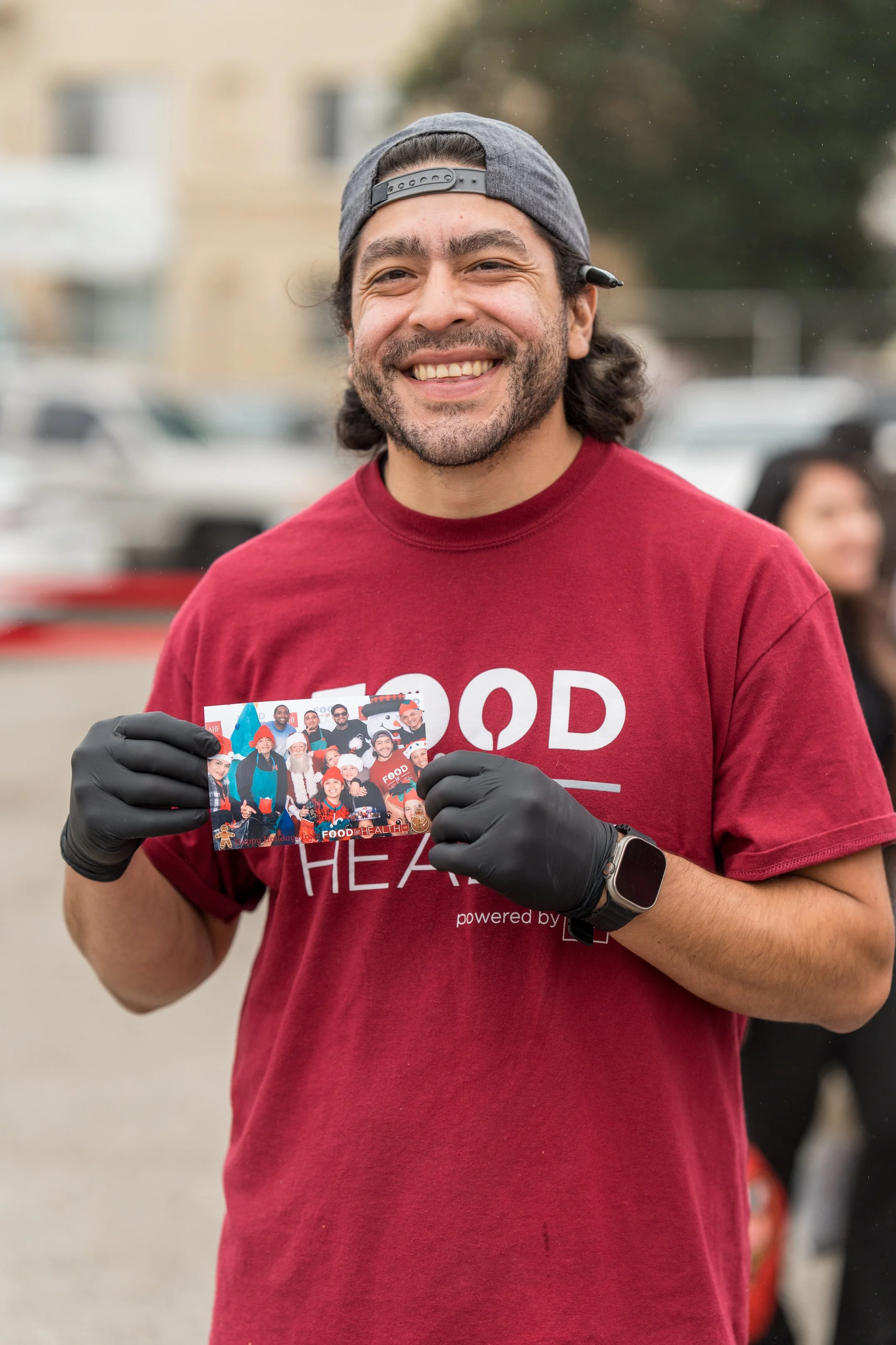 A smiling man with dark hair and a beard, wearing a gray backward cap, red T-shirt, and black gloves, holding a flyer or photo featuring a group of people in a community or charity event outdoors.