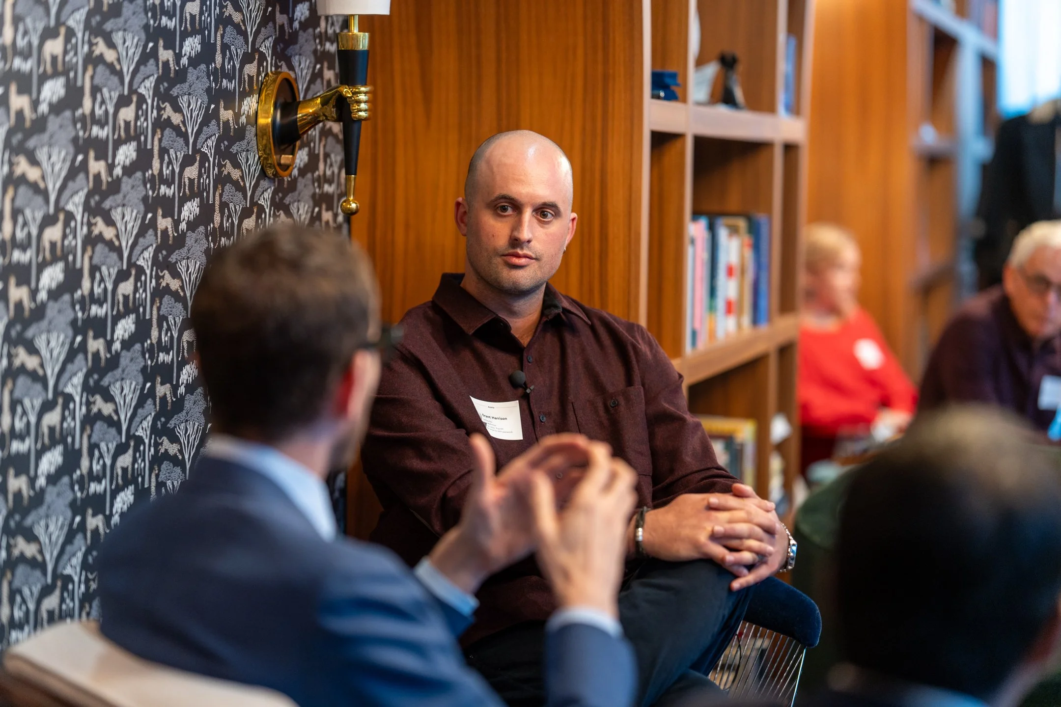 A man with a shaved head and a dark shirt is sitting at a panel discussion or meeting, surrounded by other participants. The setting has wooden shelves and decorative wallpaper with white animals and trees. People are talking and listening, with some blurred in the background.