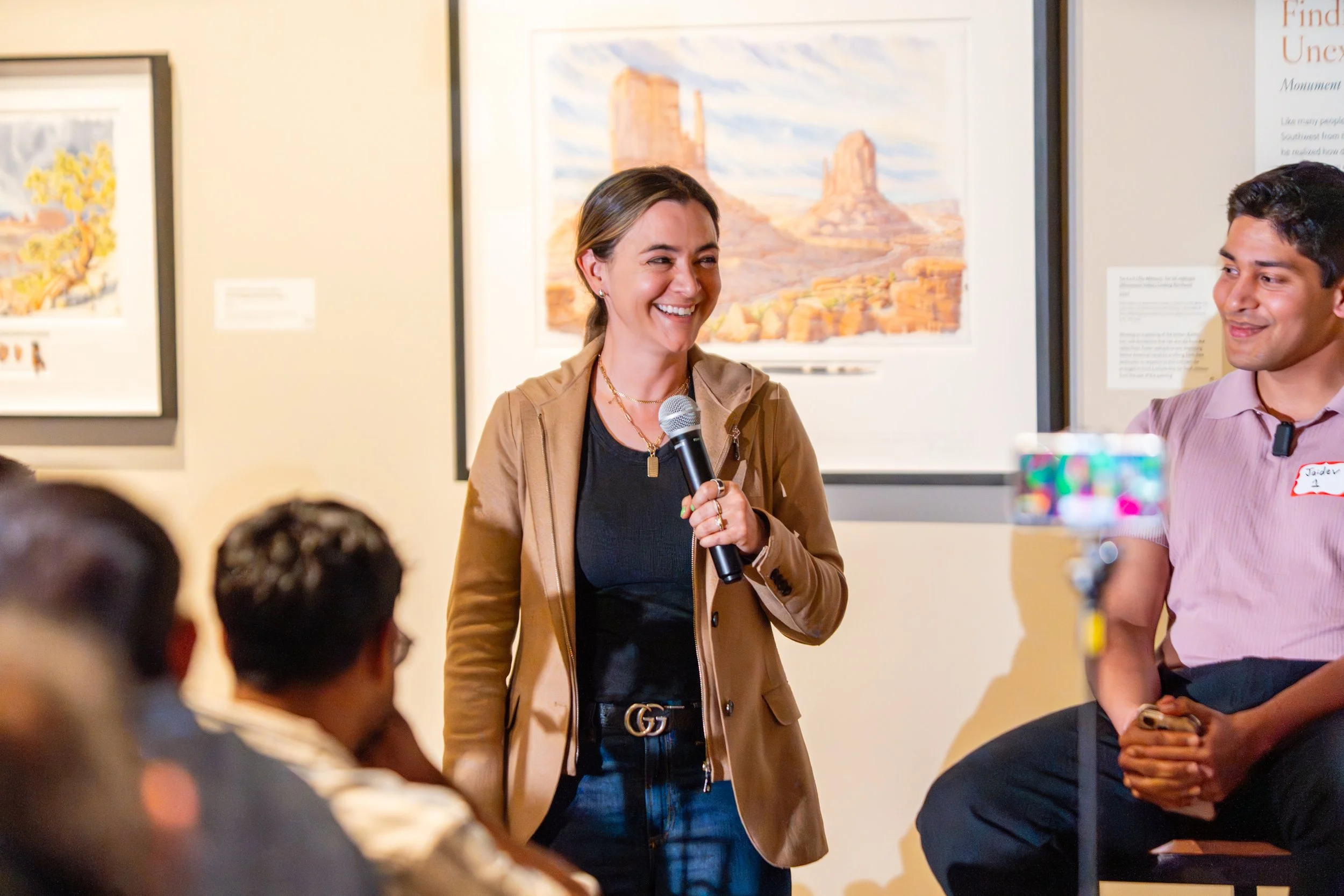 A woman holding a microphone speaking to an audience at an art gallery or museum. People sit in front of her, with framed artwork on the wall behind her.