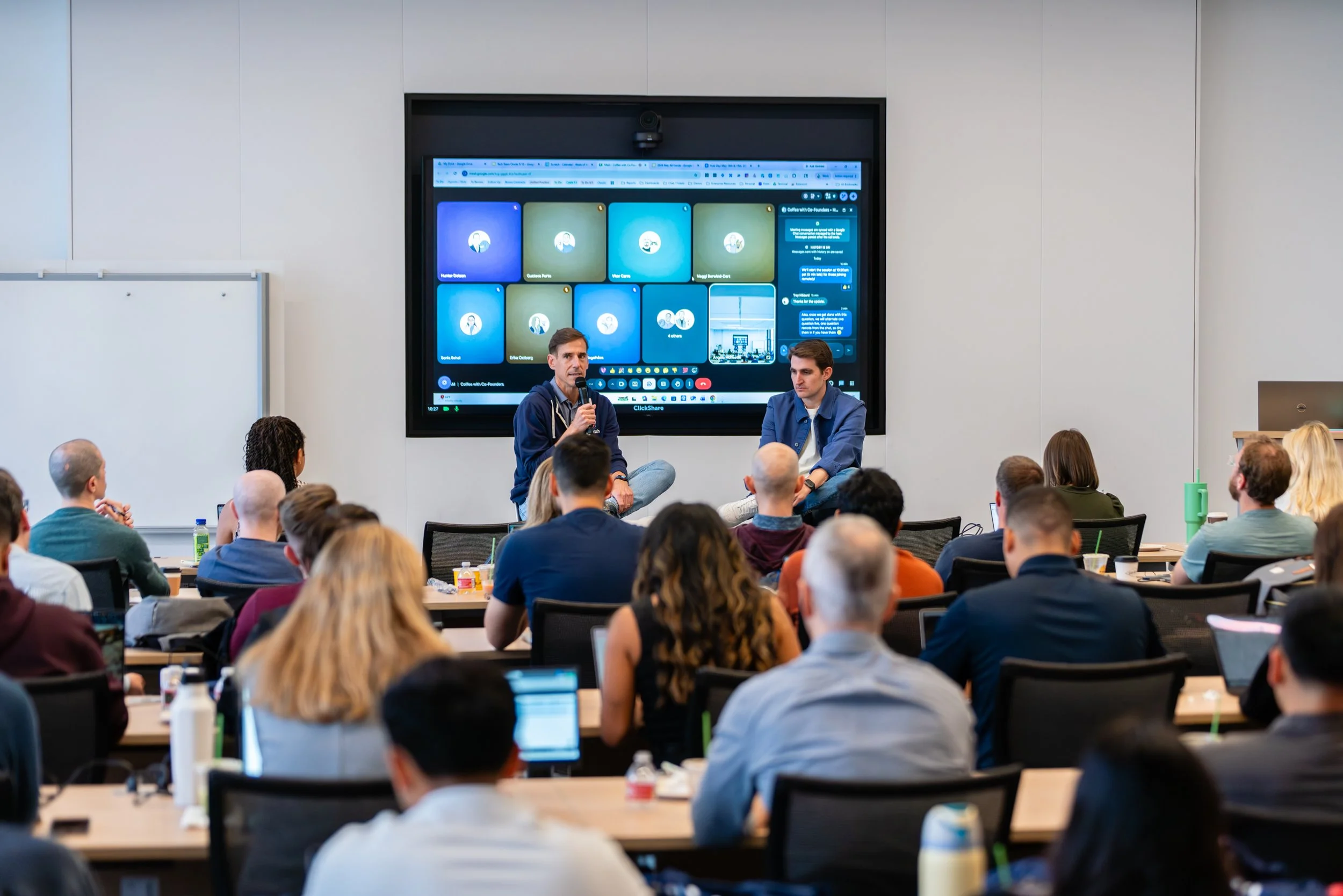A conference room filled with people attending a presentation. A man is speaking into a microphone at the front, sitting next to another man. A large screen behind them displays multiple online video call windows and chat messages. Attendees are seated at tables with laptops, drinks, and notebooks.