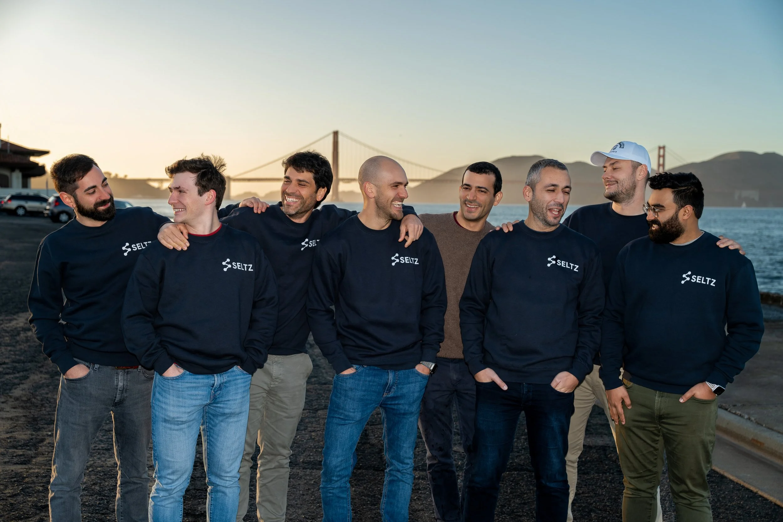 A group of eight men standing close together with arms around each other's shoulders, smiling, outdoors near water with the Golden Gate Bridge in the background.