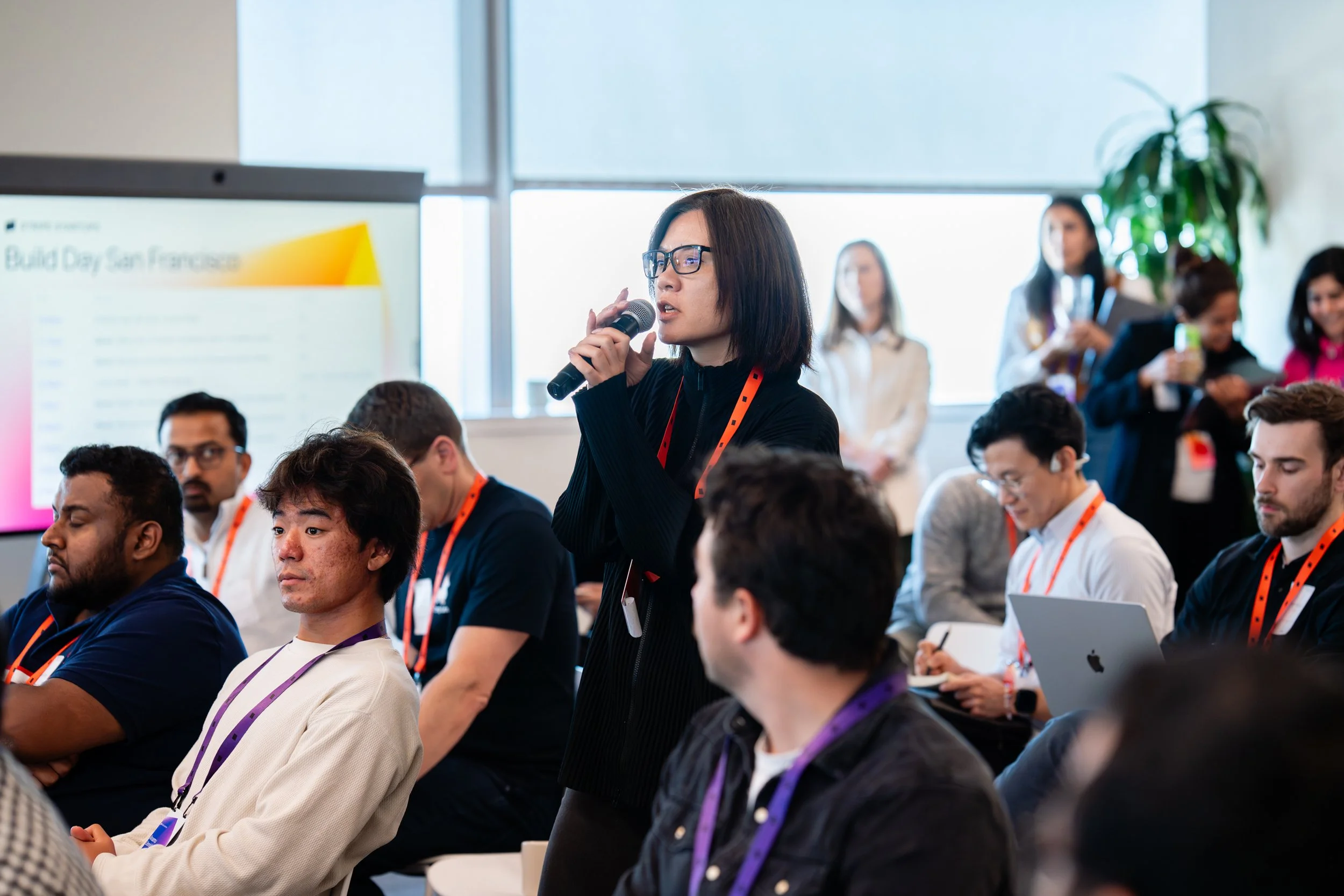 A man holding a microphone and sitting on a chair in front of an audience in a conference room featuring a large digital screen displaying a video call with multiple participants.