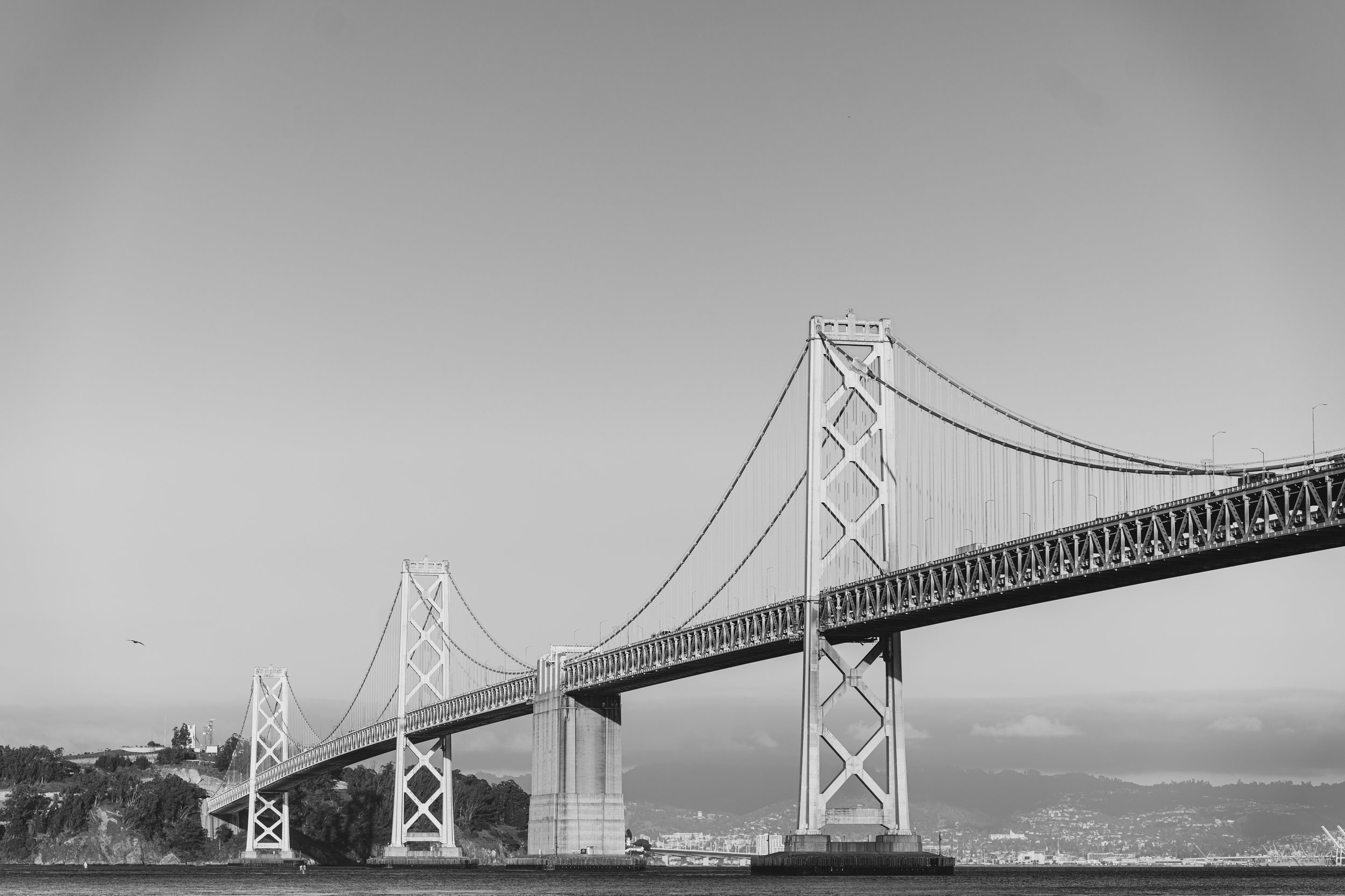Black and white photo of a large suspension bridge spanning over a body of water with hills or mountains in the background.