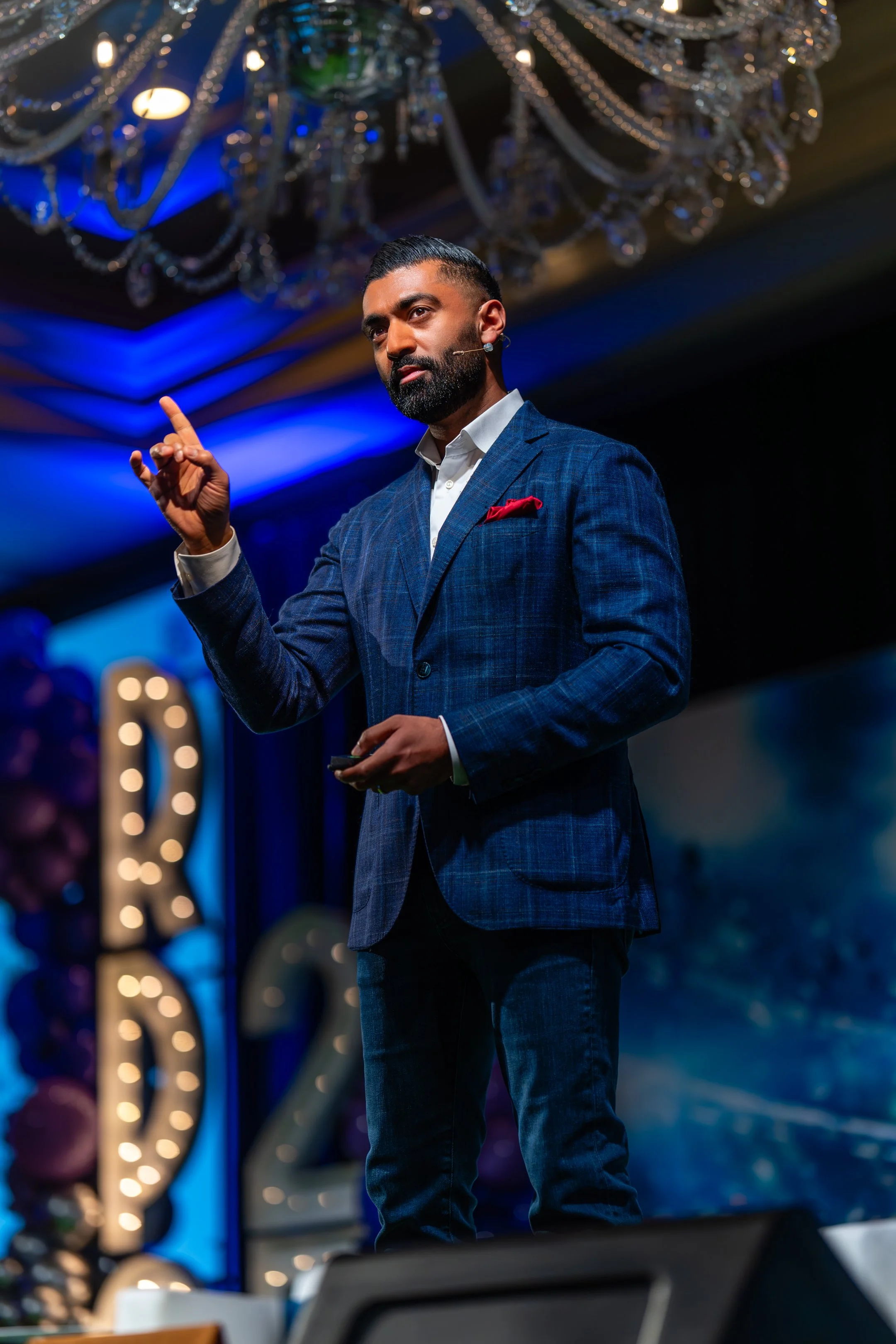 A man in a blue checkered suit giving a presentation or speech on stage with a microphone headset. There is a decorative chandelier hanging from the ceiling and a blurred illuminated sign that reads 'R' in the background.