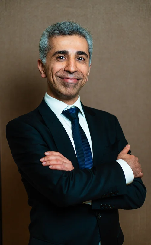 Team headshot of a man in a black suit with arms crossed on a brown backdrop