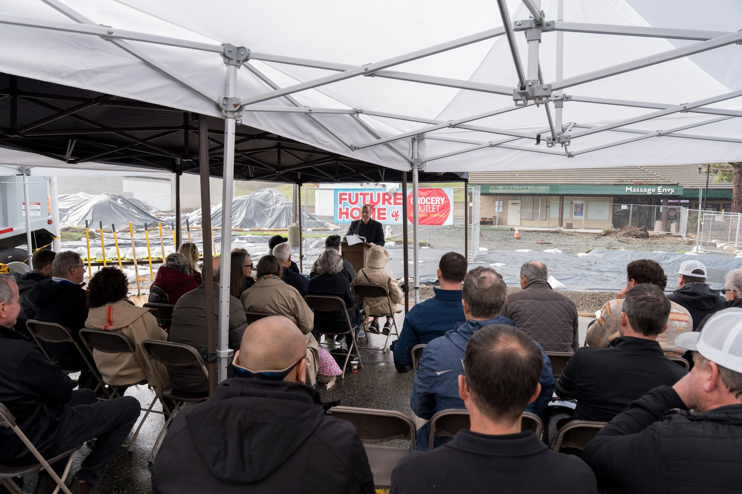 A man giving a presentation to a seated audience under a tent at a construction site, with a cloudy sky and construction signs in the background.