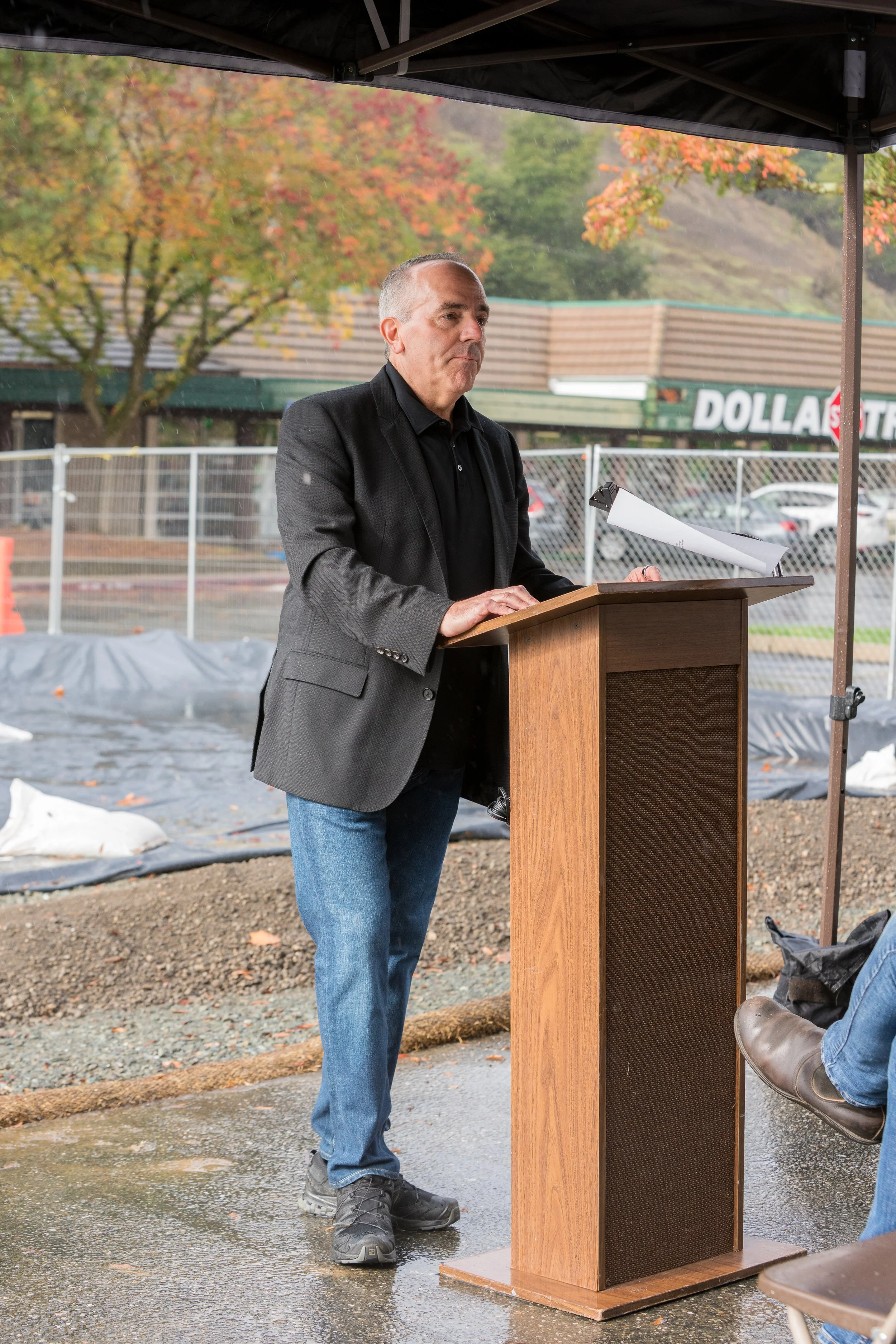 A man in a black blazer and jeans stands at a wooden podium, speaking or preparing to speak at an outdoor event under a canopy on a rainy day. Raindrops are visible, and the background features autumn-colored trees and a fenced parking lot with parked cars.