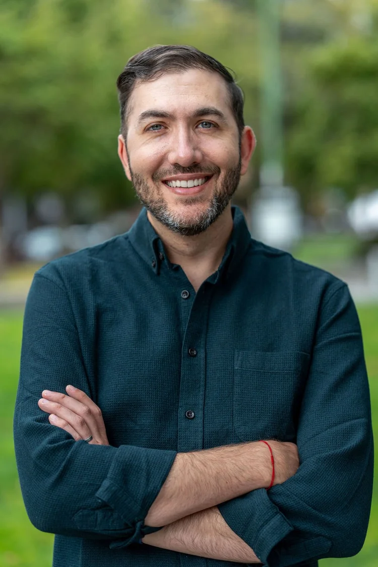 Executive portrait of a smiling man in a dark green shirt with arms crossed outdoors