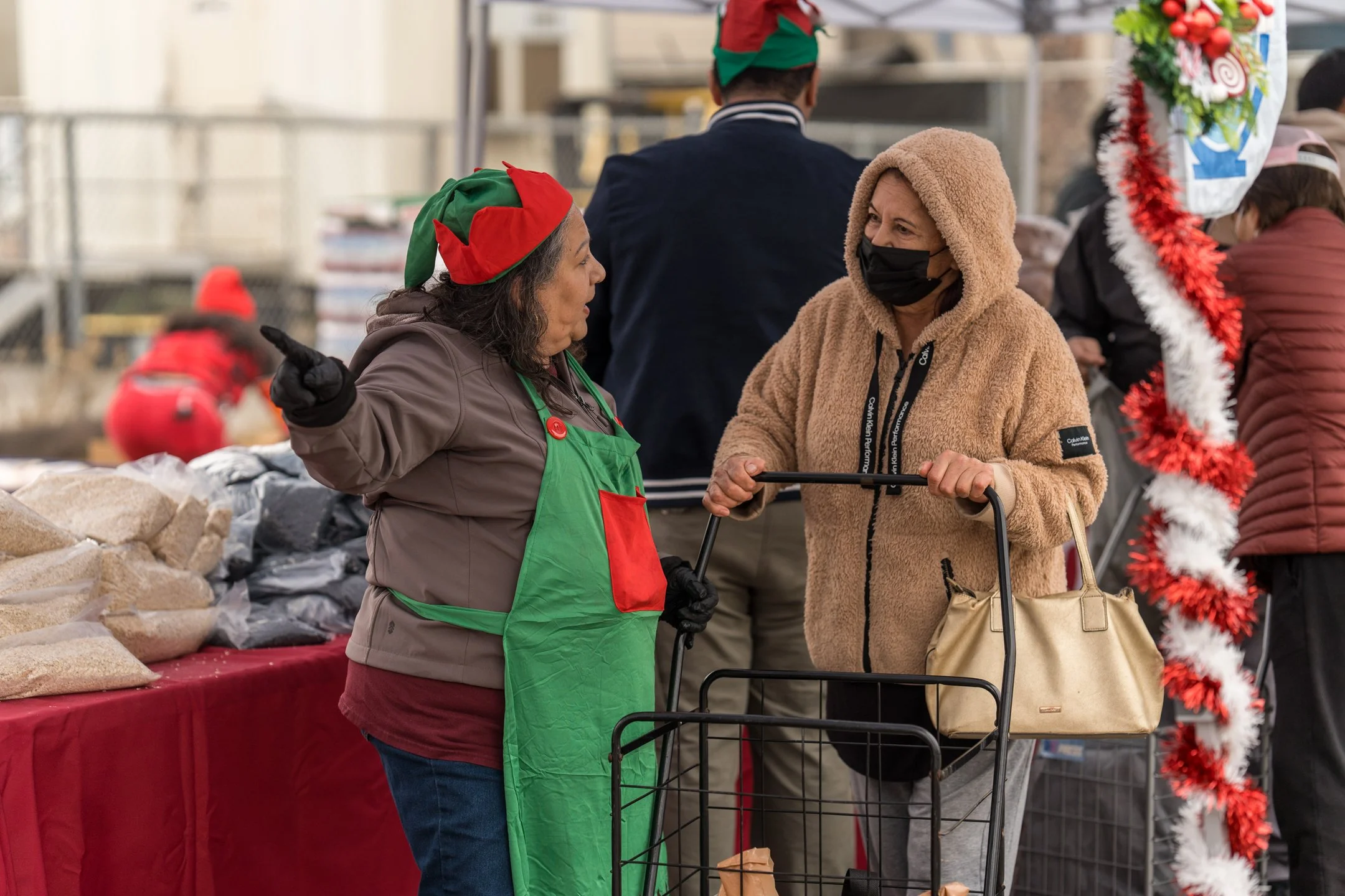 Two women, one in a green apron and festive elf hat, and the other in a beige hoodie and black face mask, converse at a holiday market stall decorated with red and white tinsel. The woman in the apron is gesturing with her finger as they talk, with bags of bread and other items on the red table.