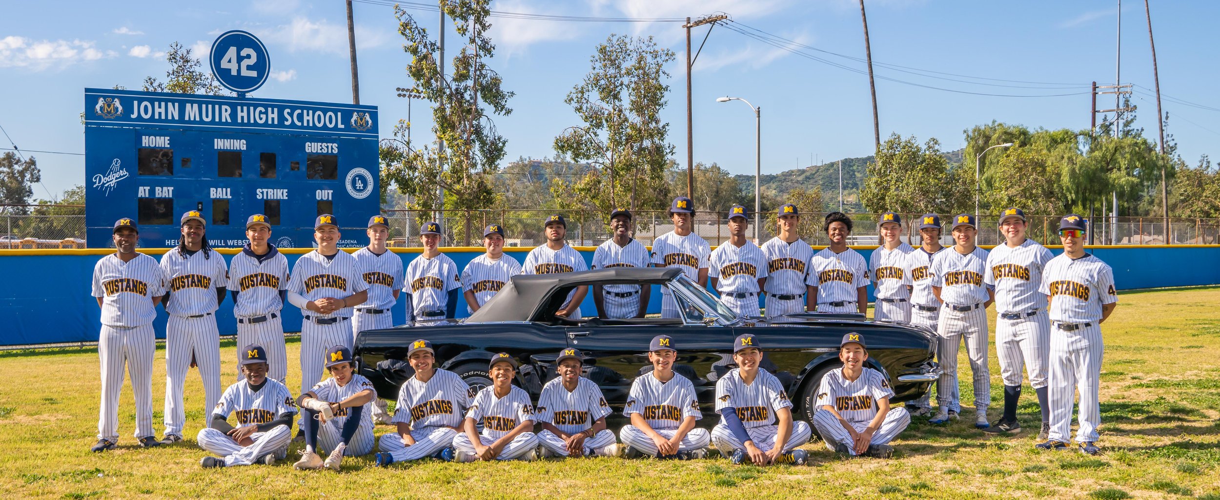 A high school baseball team called the Mustangs poses together on the field with a classic black car. The players are dressed in white pinstriped uniforms with 'Mustangs' written on the front and yellow caps with an M. They are standing and sitting i