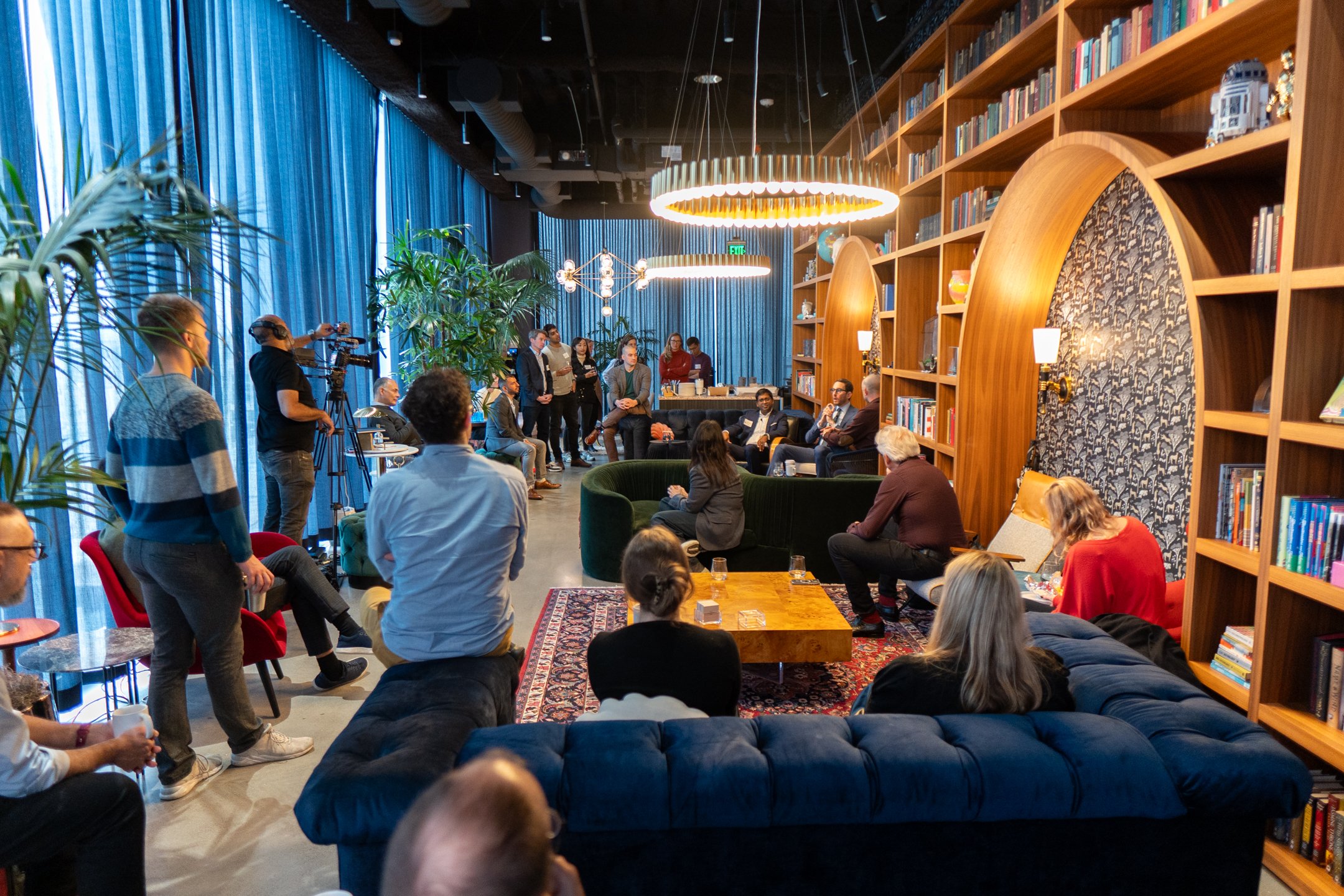 A group of people attending a panel discussion or presentation in a modern, cozy library or lounge space with blue curtains, large bookshelves, and stylish lighting fixtures.
