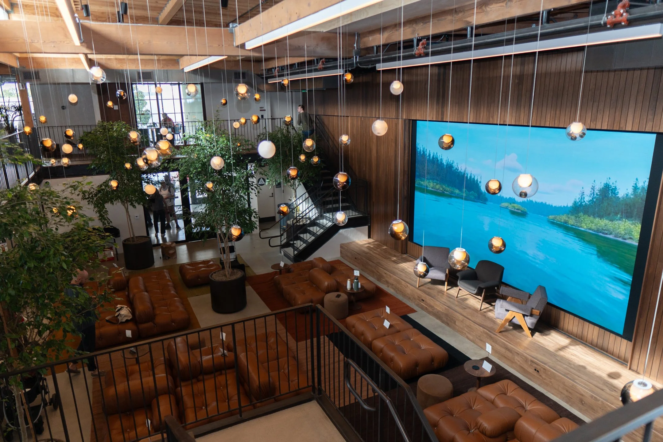 Interior view of a spacious modern building with a high ceiling and wooden beams. There are hanging decorative spherical lights, a large digital screen displaying a nature scene, and multiple seating areas with brown leather sofas and chairs. Indoor 