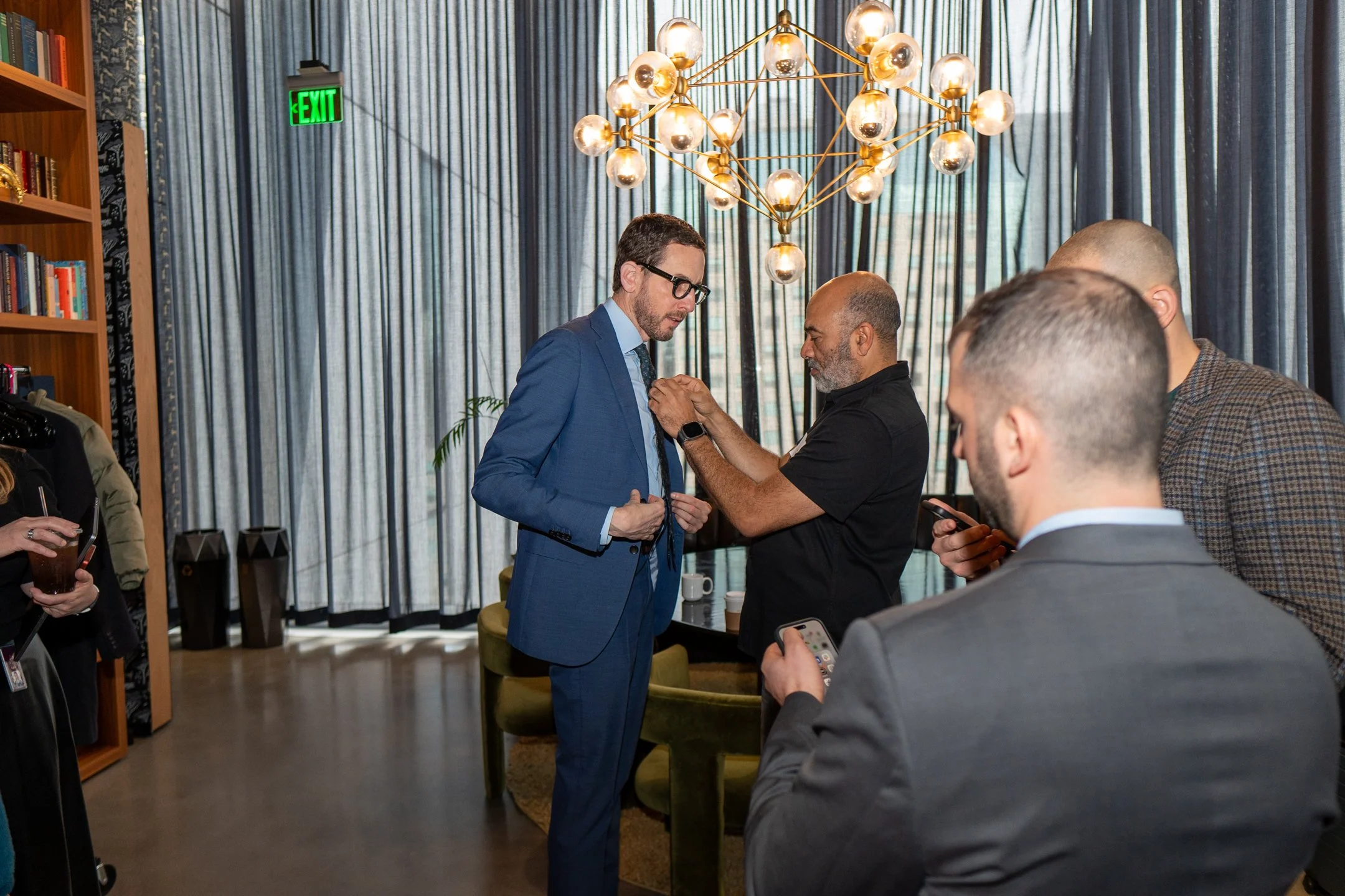 A man in a blue suit, glasses, and a beard is receiving a pin or medal from a woman in a black shirt with gray hair and a beard, in a room with large blue curtains and a modern chandelier. Several people are gathered around, some looking at their phones, and a bookshelf is visible on the left side.