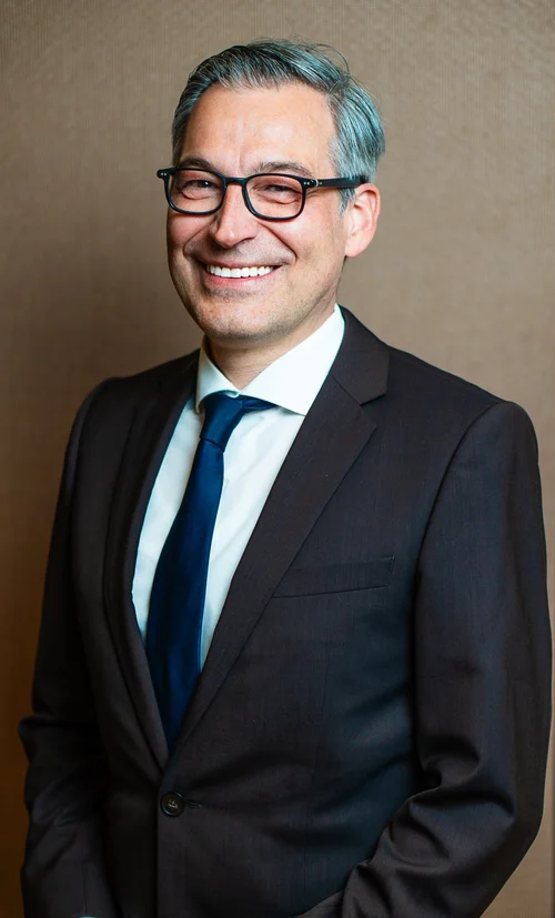 Team headshot of a man in a dark suit and glasses on a brown backdrop