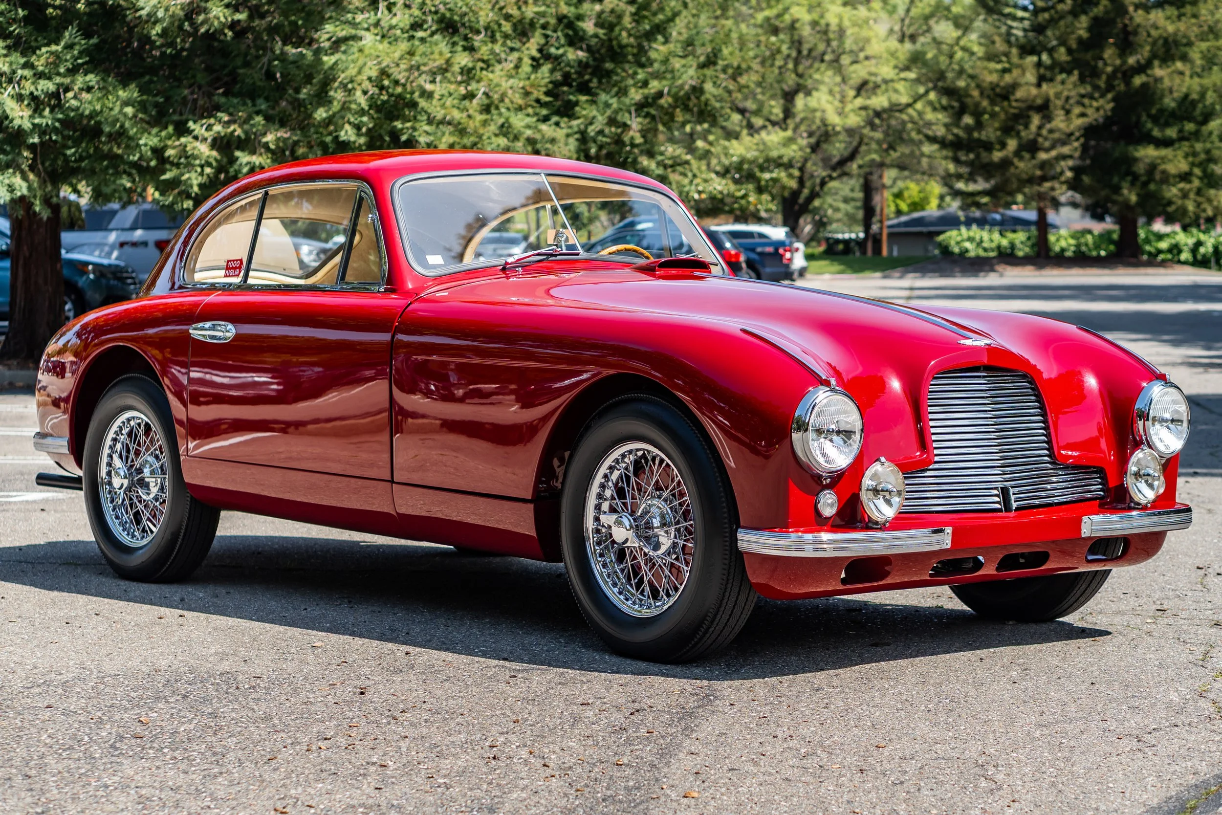 A shiny red vintage sports car with chrome wire wheels parked in a parking lot under trees.