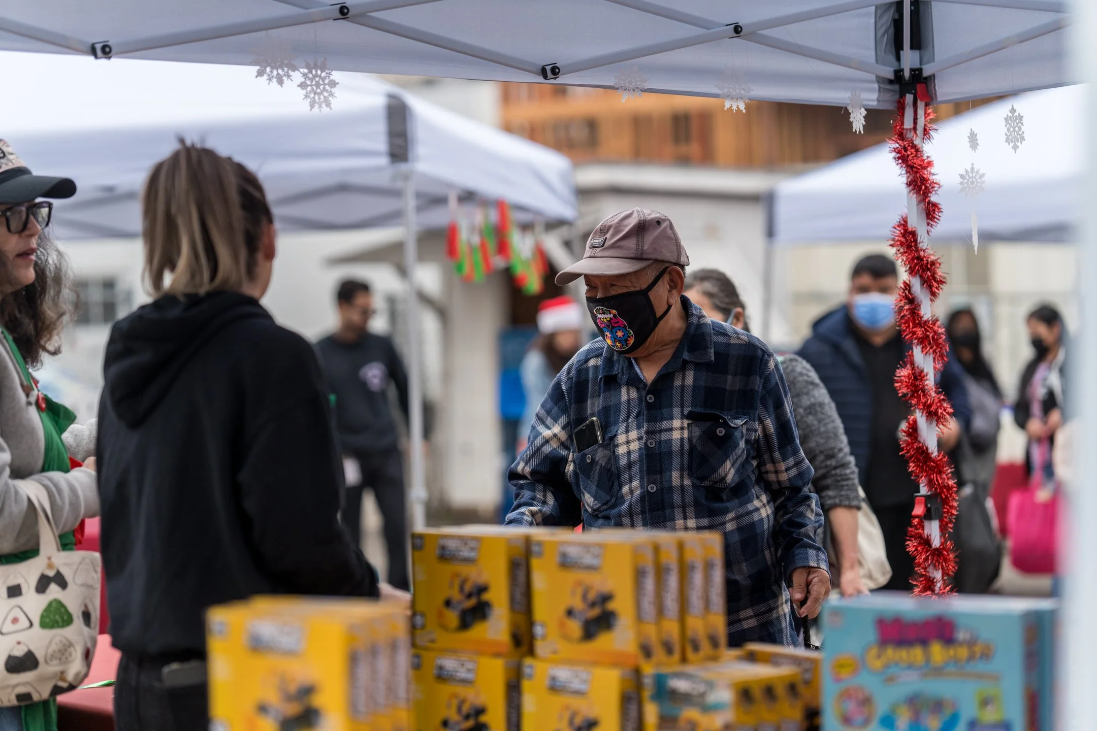 People shopping at an outdoor holiday market, wearing masks, with Christmas decorations hanging from tents and holiday-themed products on display.