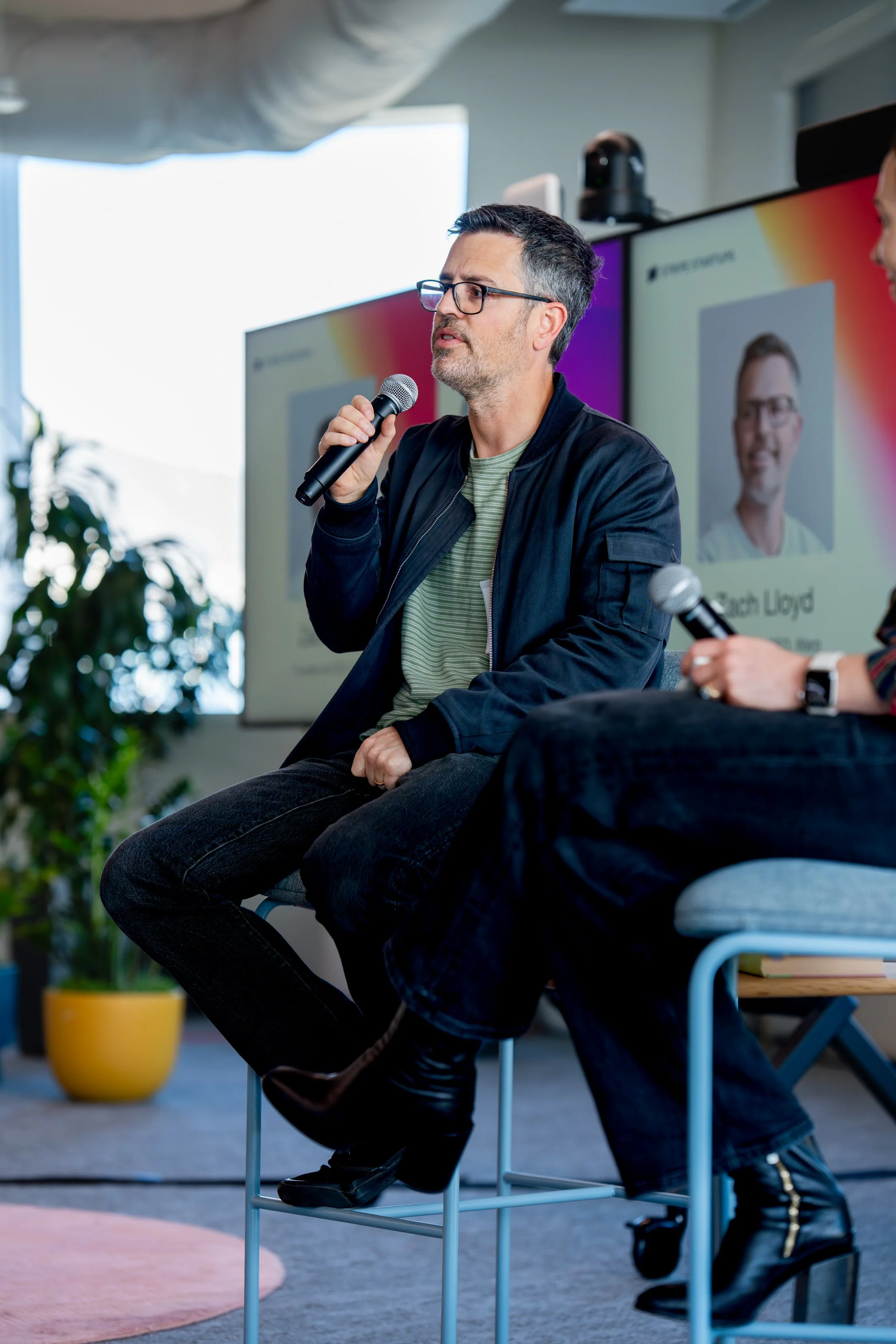A man with glasses and a beard, wearing a black jacket and green t-shirt, holds a microphone and speaks during a panel discussion or presentation. There is a large screen behind him with a projection of his face and name. Another person is partially visible holding a microphone. The setting appears to be an indoor event space with natural light, plants, and modern furniture.