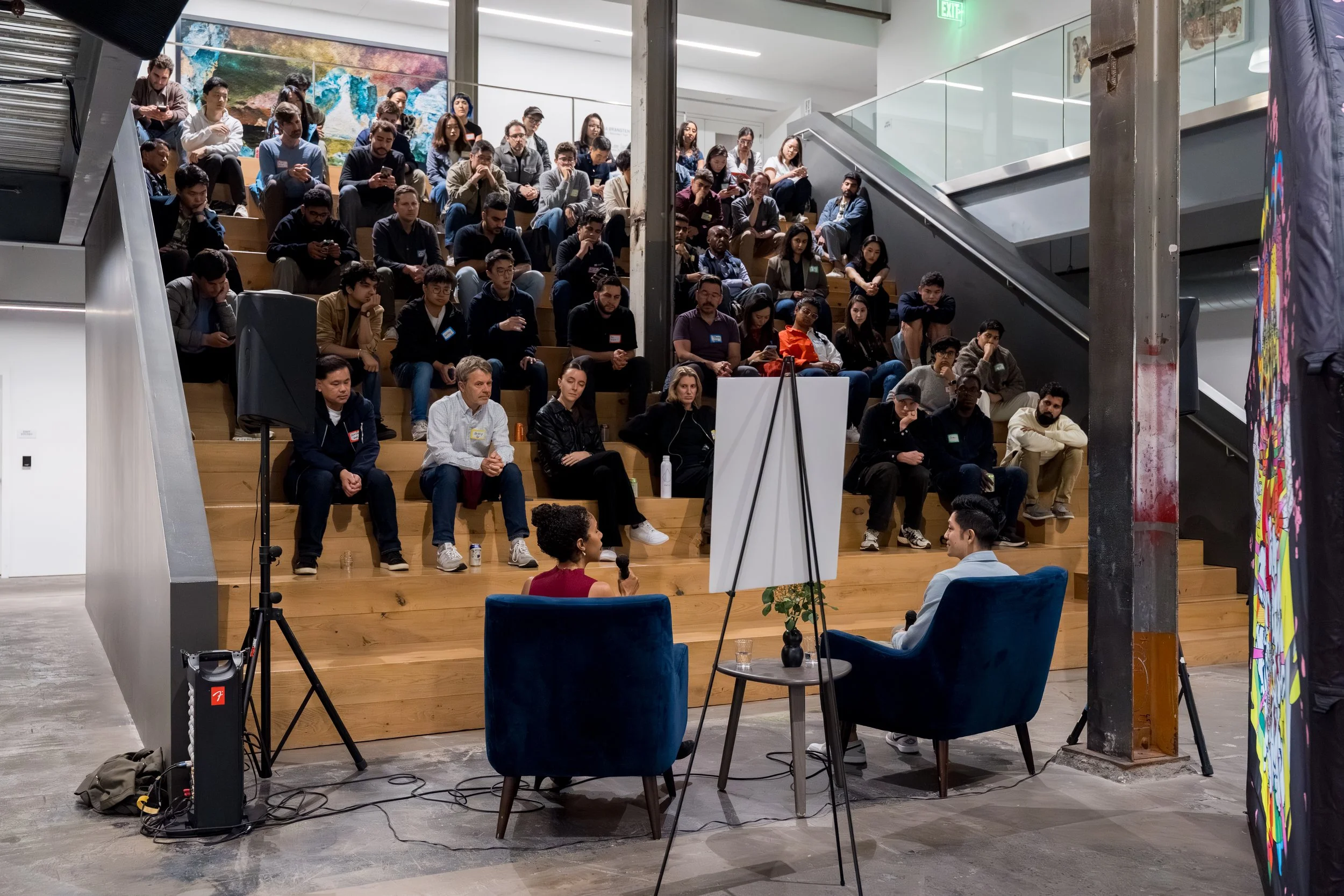 Audience seated on wooden tiered steps attending a presentation or panel discussion in an indoor setting, with two people speaking at a table in front.