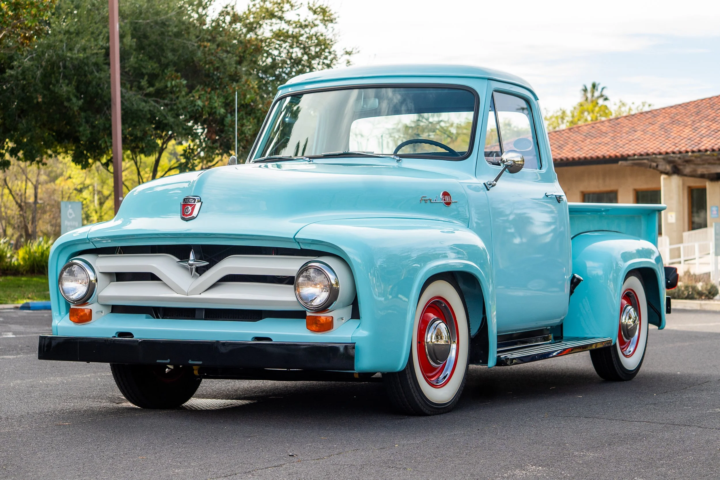 A vintage light blue pickup truck parked outdoors in front of a building with a red-tiled roof and surrounded by green trees.