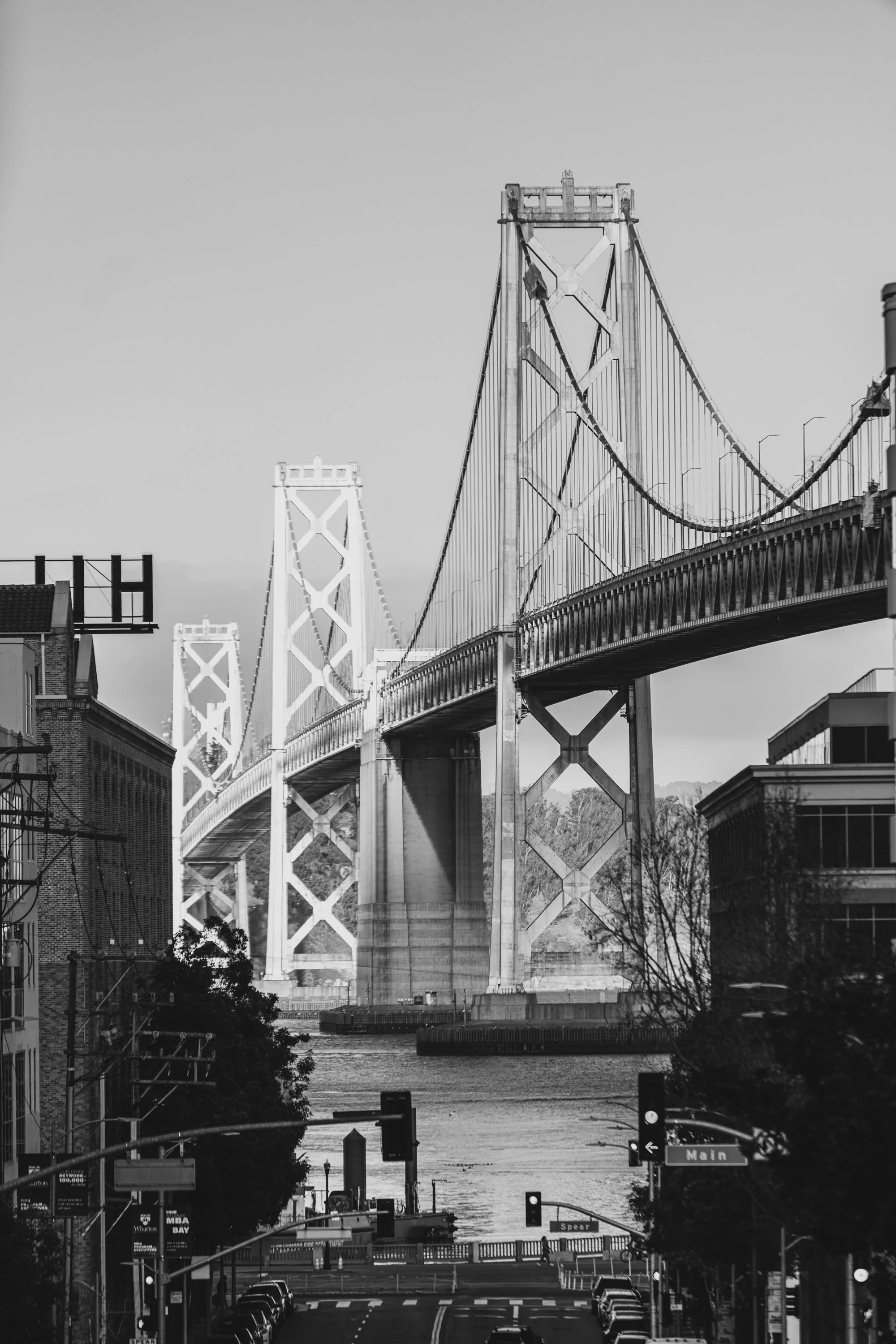 Black and white photo of a bridge spanning a river, seen from below street level with buildings lining the streets on each side. Traffic lights and street signs are visible, with the main road leading toward the bridge.
