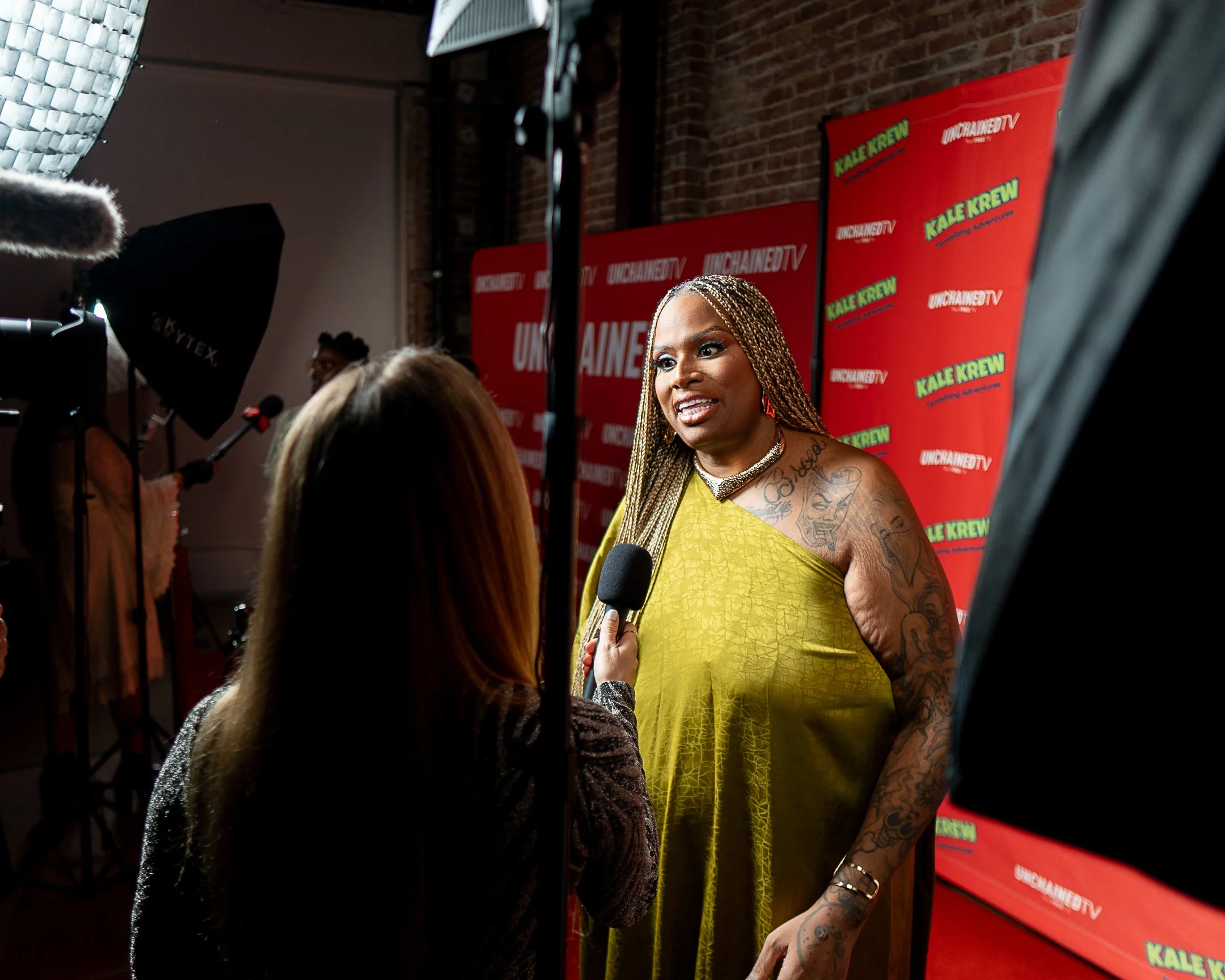 Woman with braided hair, tattoos, and a yellow dress speaking into a microphone during an interview, with a red 'Unchained TV' backdrop featuring green 'Kale Krew' logos in the background.