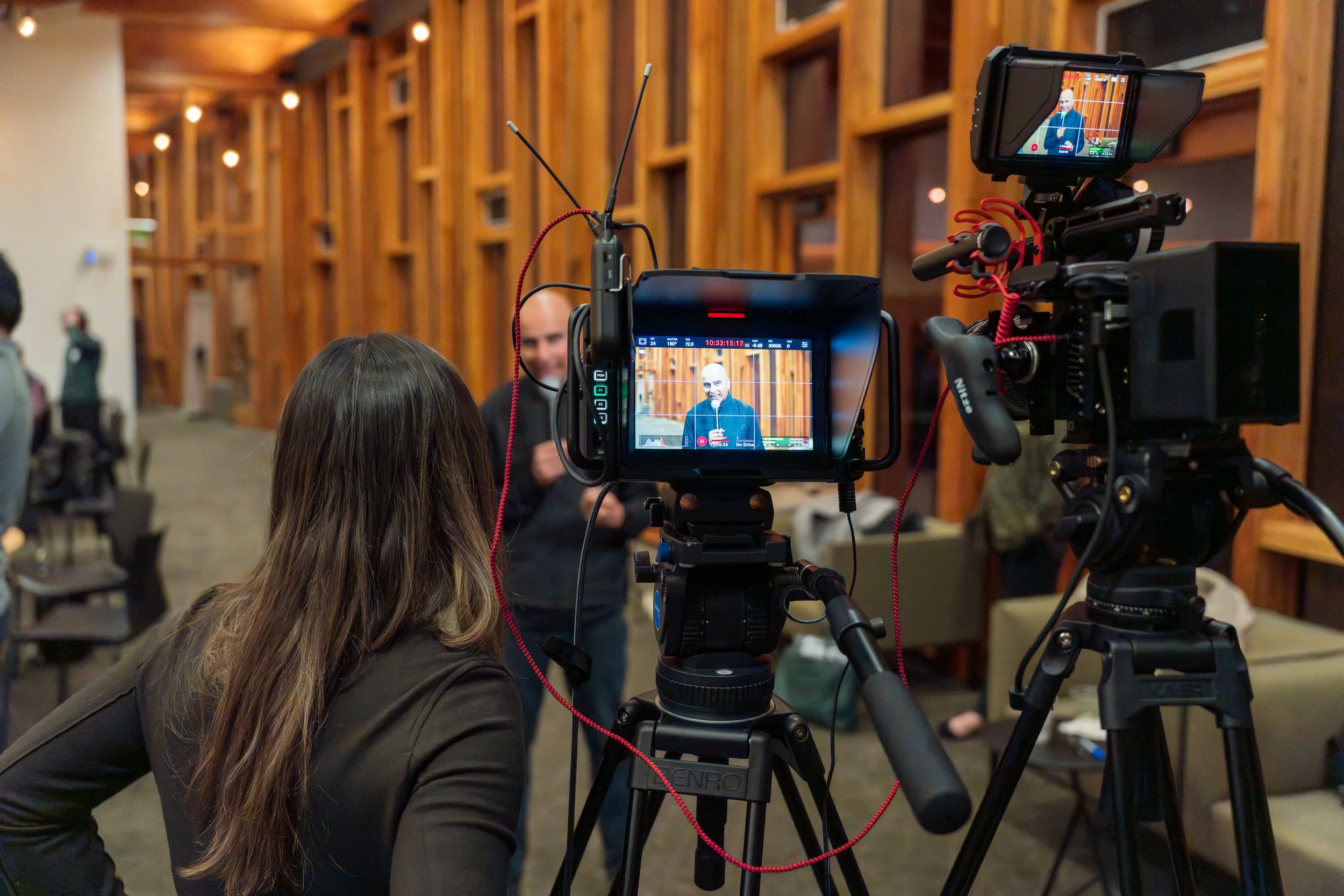 A person with long brown hair wearing a black jacket is operating a video camera on a tripod, recording a man speaking in front of a wooden wall with geometric patterning. The man being recorded is in focus on the camera's viewfinder, wearing glasses and a dark blazer, holding a microphone. There are people and chairs in the background of the indoor setting.
