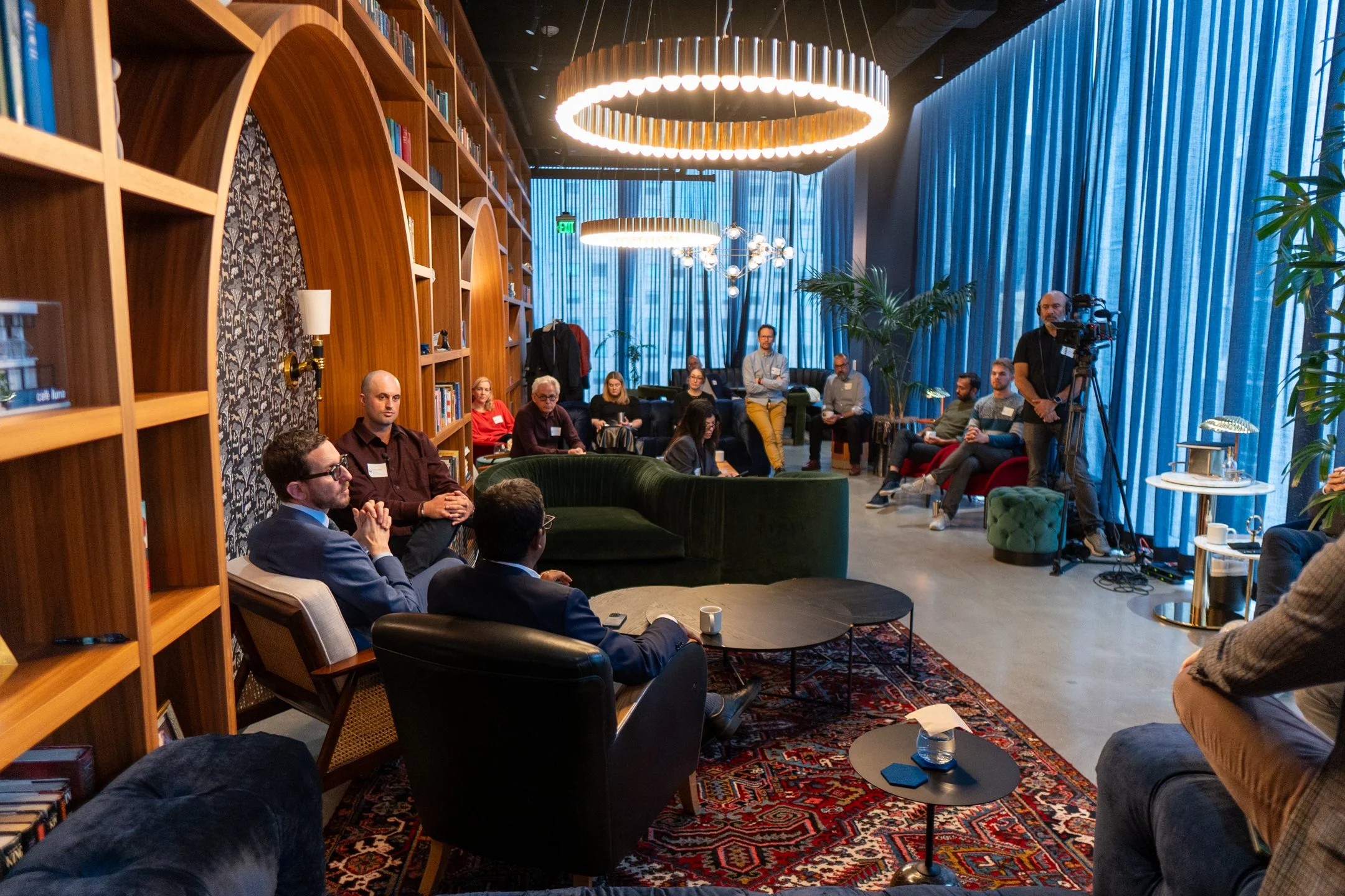 A group of people in a cozy, well-lit library or lounge area, some seated and some standing, engaging in a discussion or presentation. There are bookshelves, large indoor plants, and modern lighting fixtures.