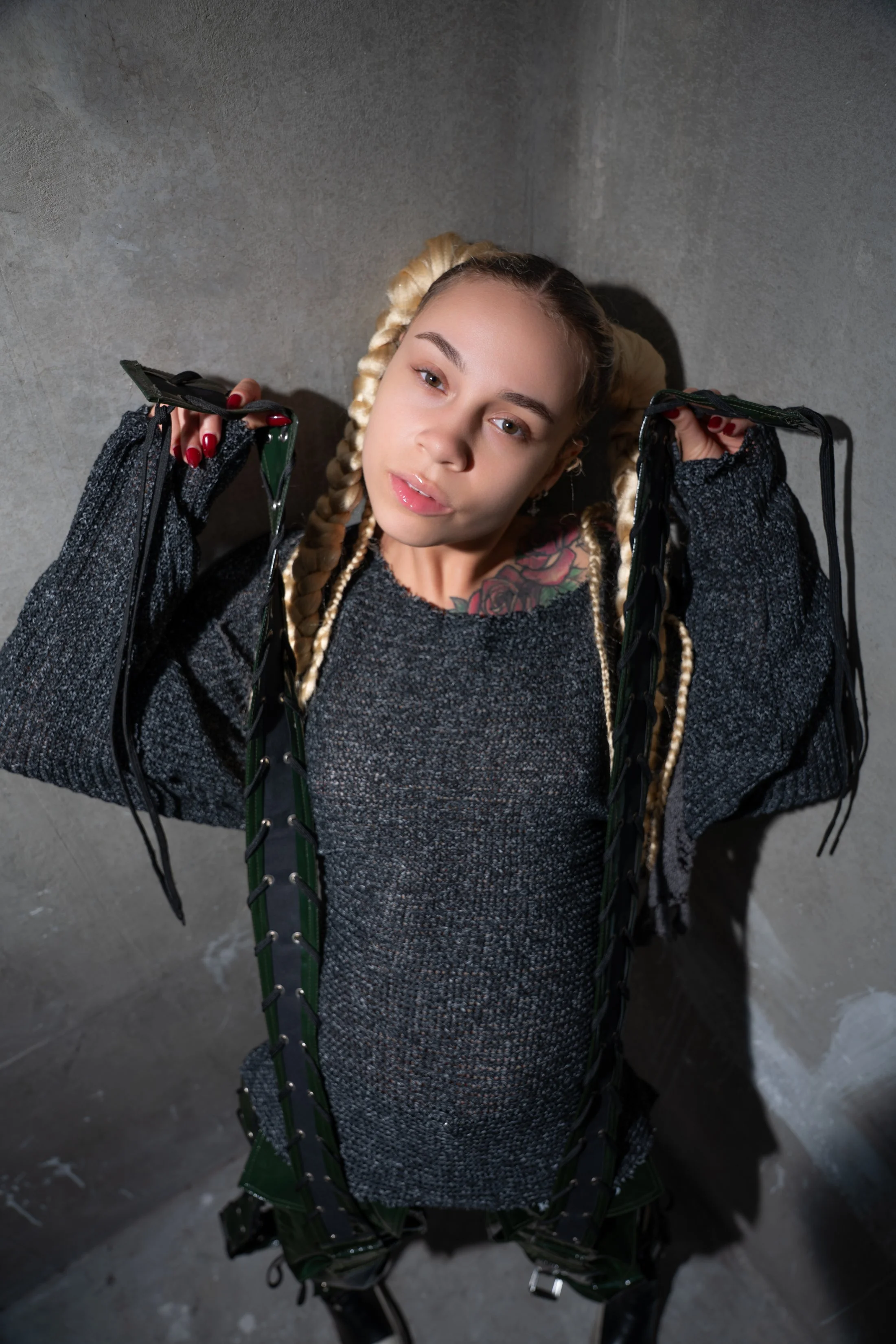A young woman with long blonde braided hair and tattoos on her chest wears a dark sweater and holds open a black and green harness, looking into the camera against a concrete wall.