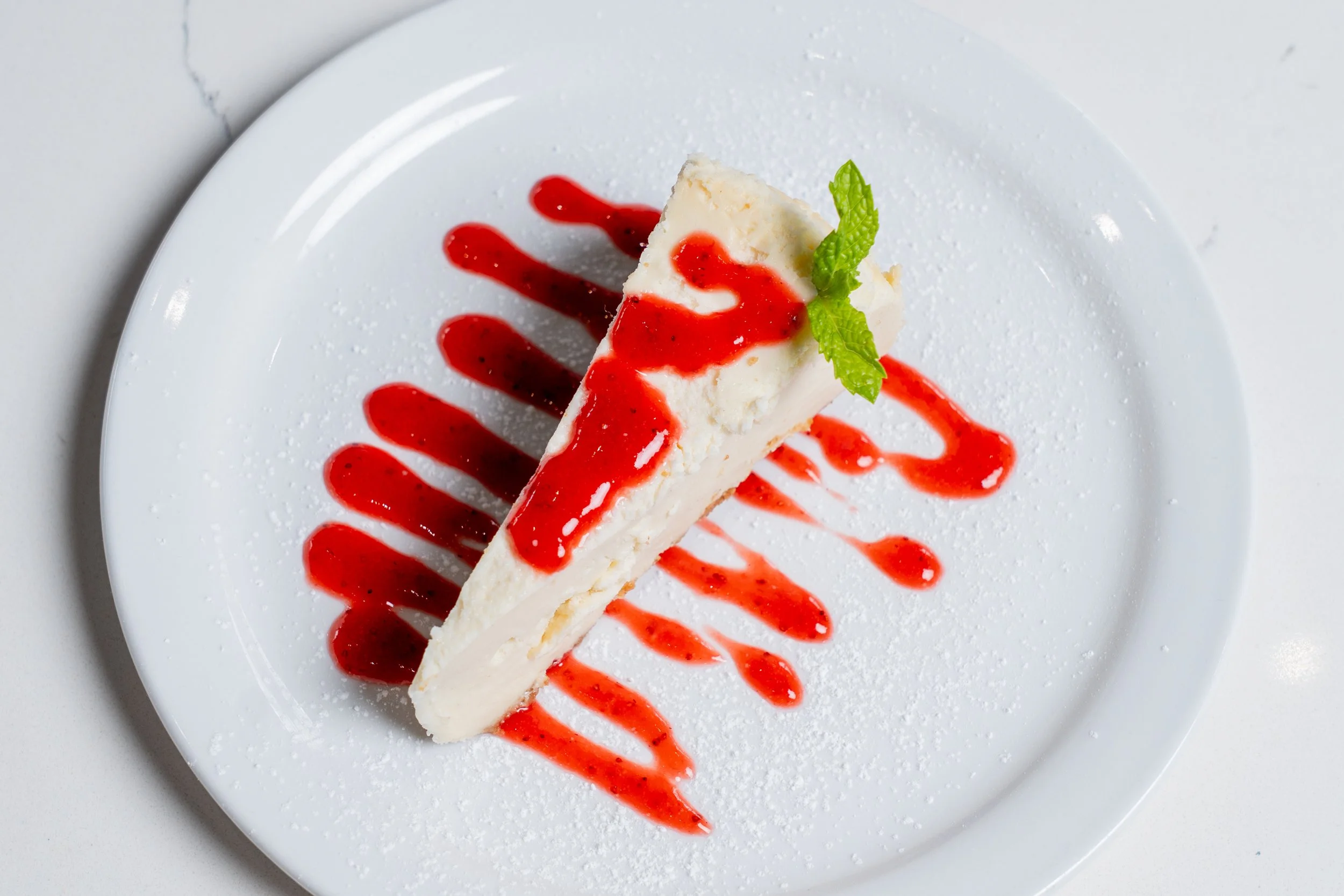 Slice of cheesecake on a white plate drizzled with red berry sauce and garnished with a green mint leaf.